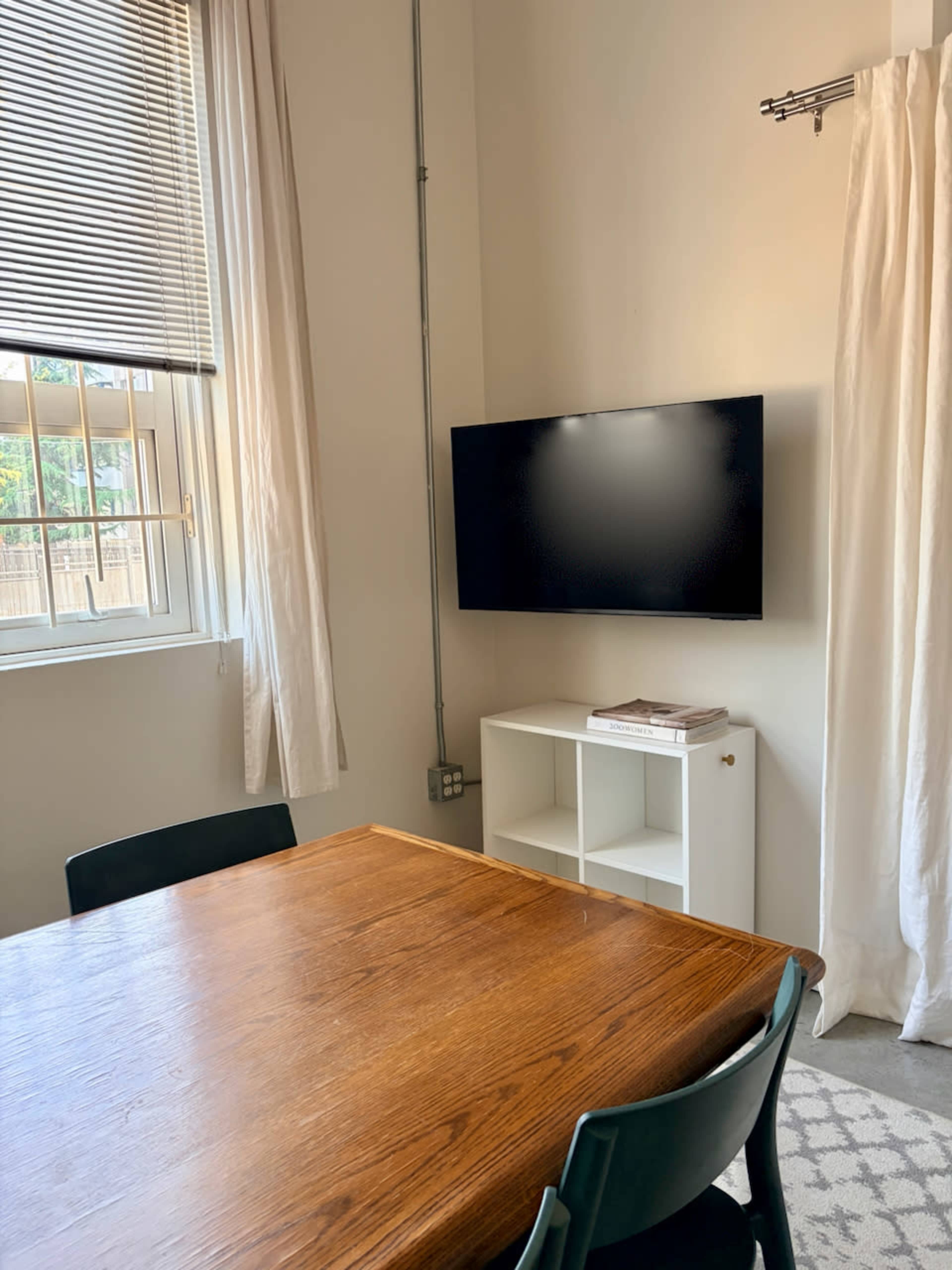 A wooden dining table with black chairs is positioned in a well-lit room featuring a television mounted on the wall and a white shelving unit nearby.