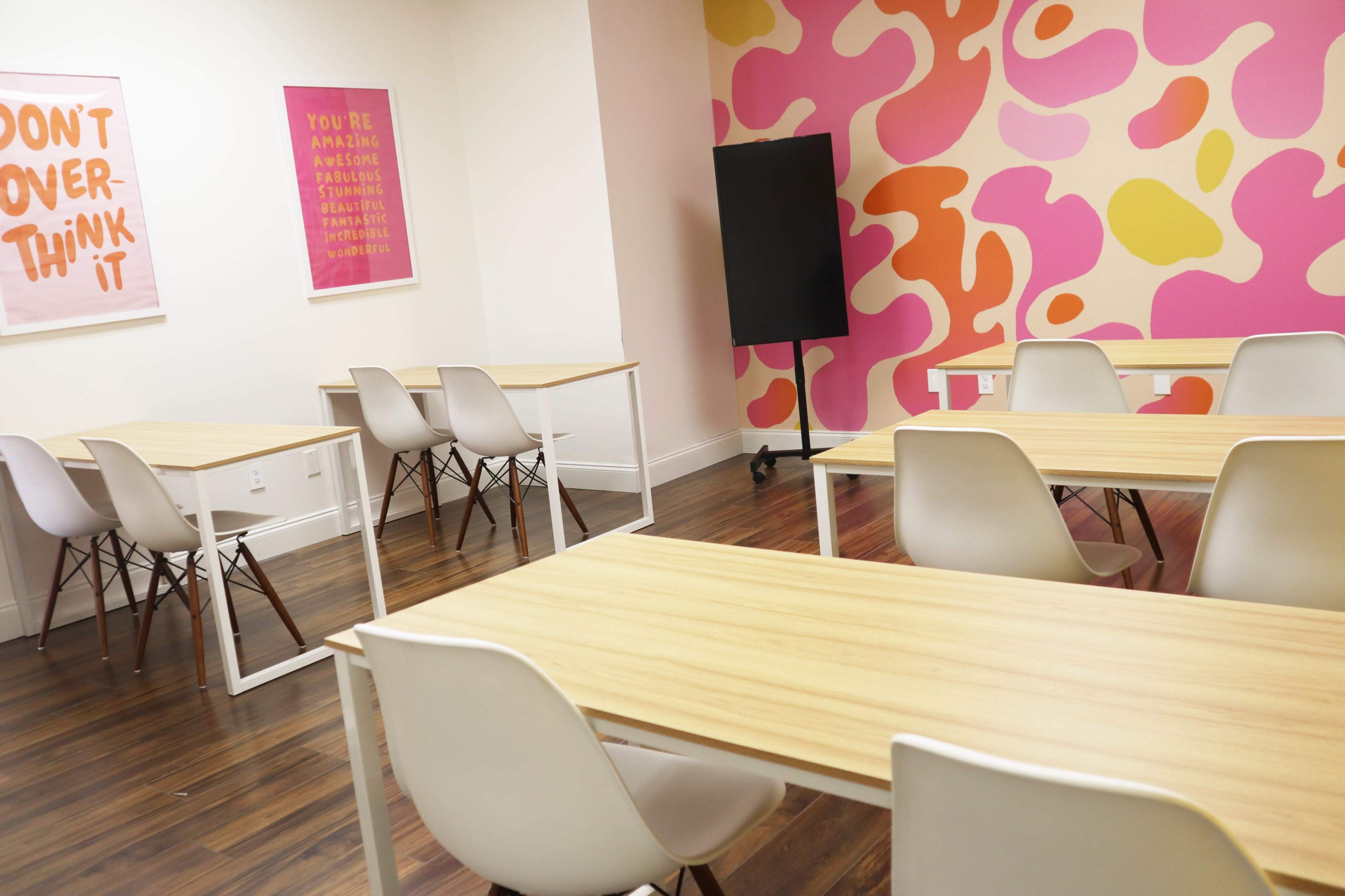 A brightly colored classroom with several tables and chairs arranged around a feature wall adorned with abstract pink and yellow patterns.