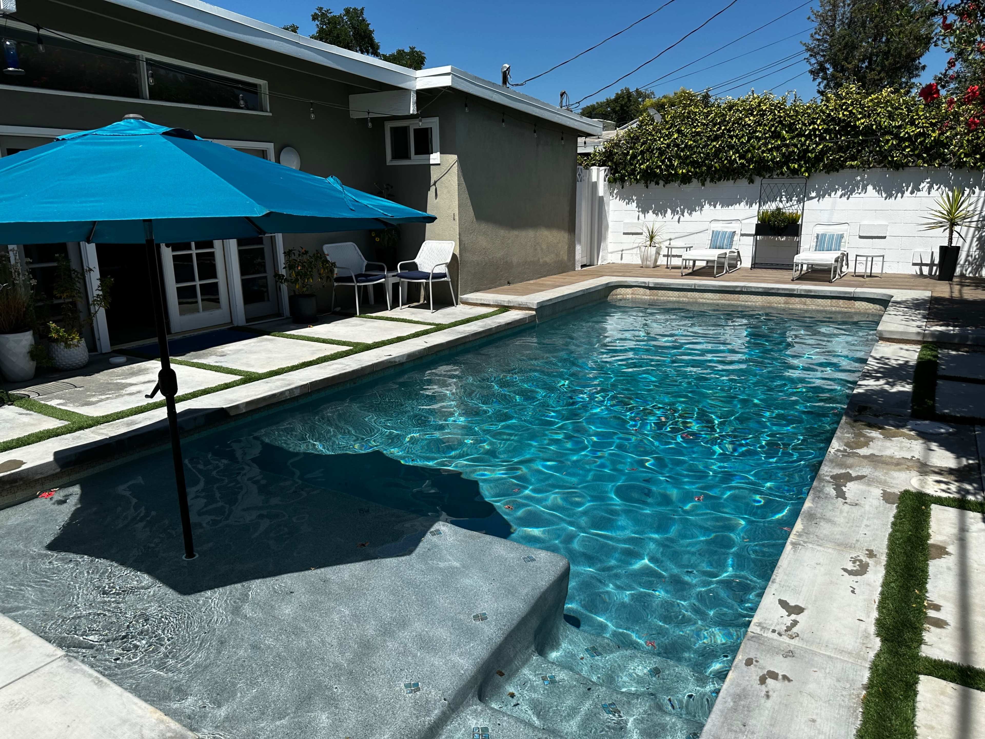 The image shows a clear swimming pool surrounded by a stone deck, an umbrella, and a few lounge chairs against a backdrop of greenery and a building.