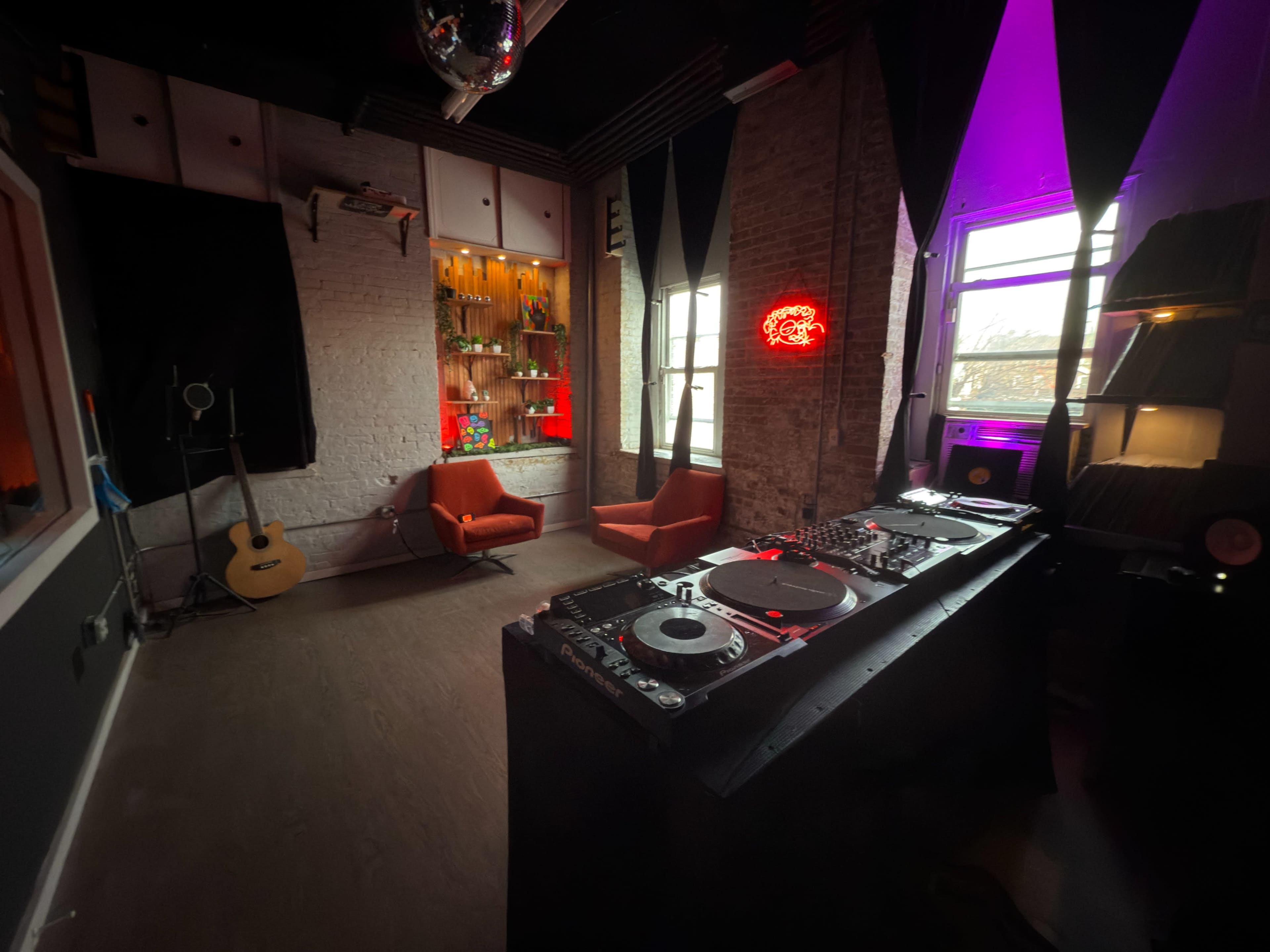 The image shows a dimly lit room equipped with DJ decks, a guitar, two orange chairs, and a bar with illuminated shelves.