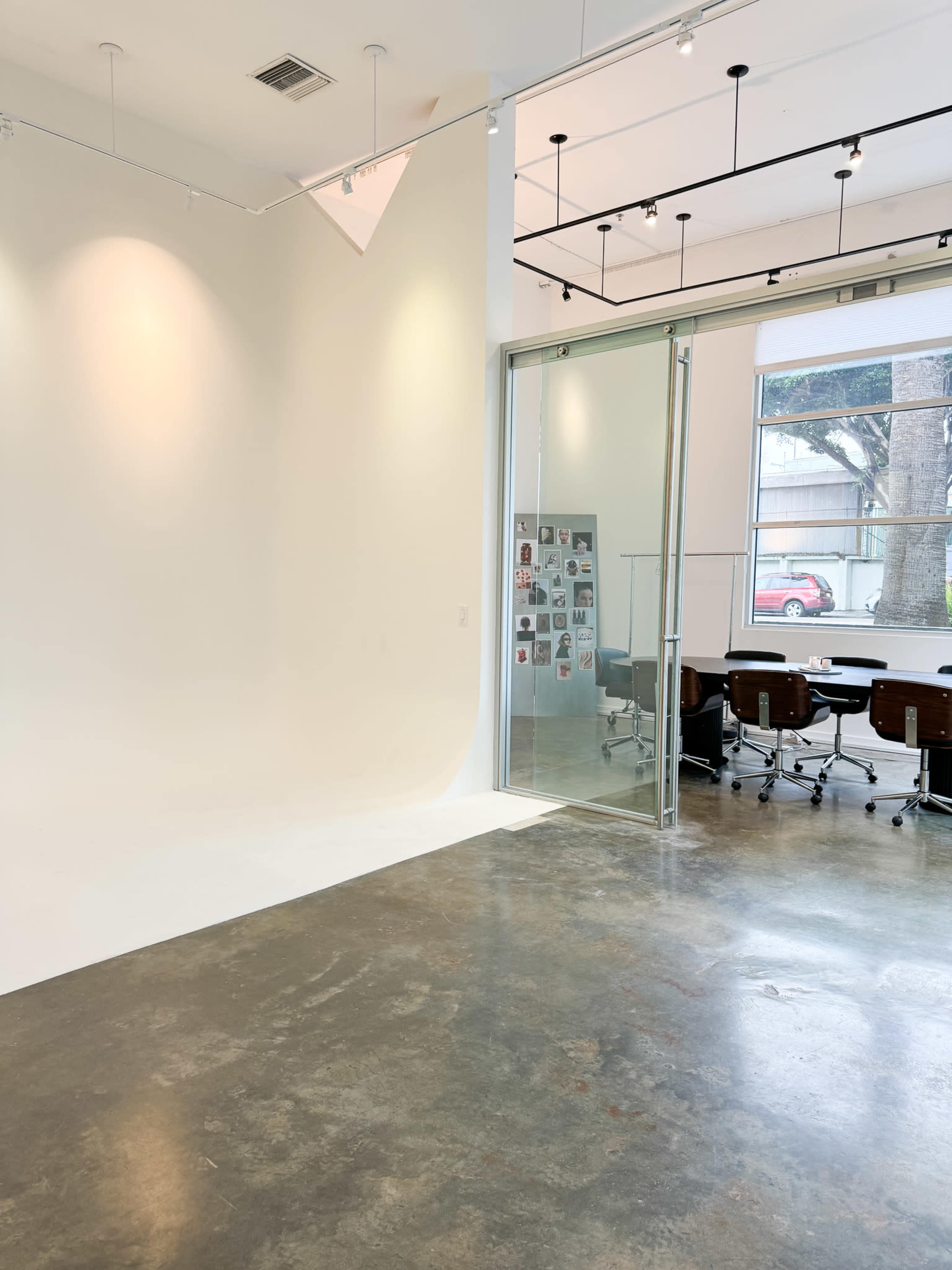 The image shows a modern office space featuring a blank white wall, a glass door with a collage on the adjacent wall, and a conference table with chairs in the background.