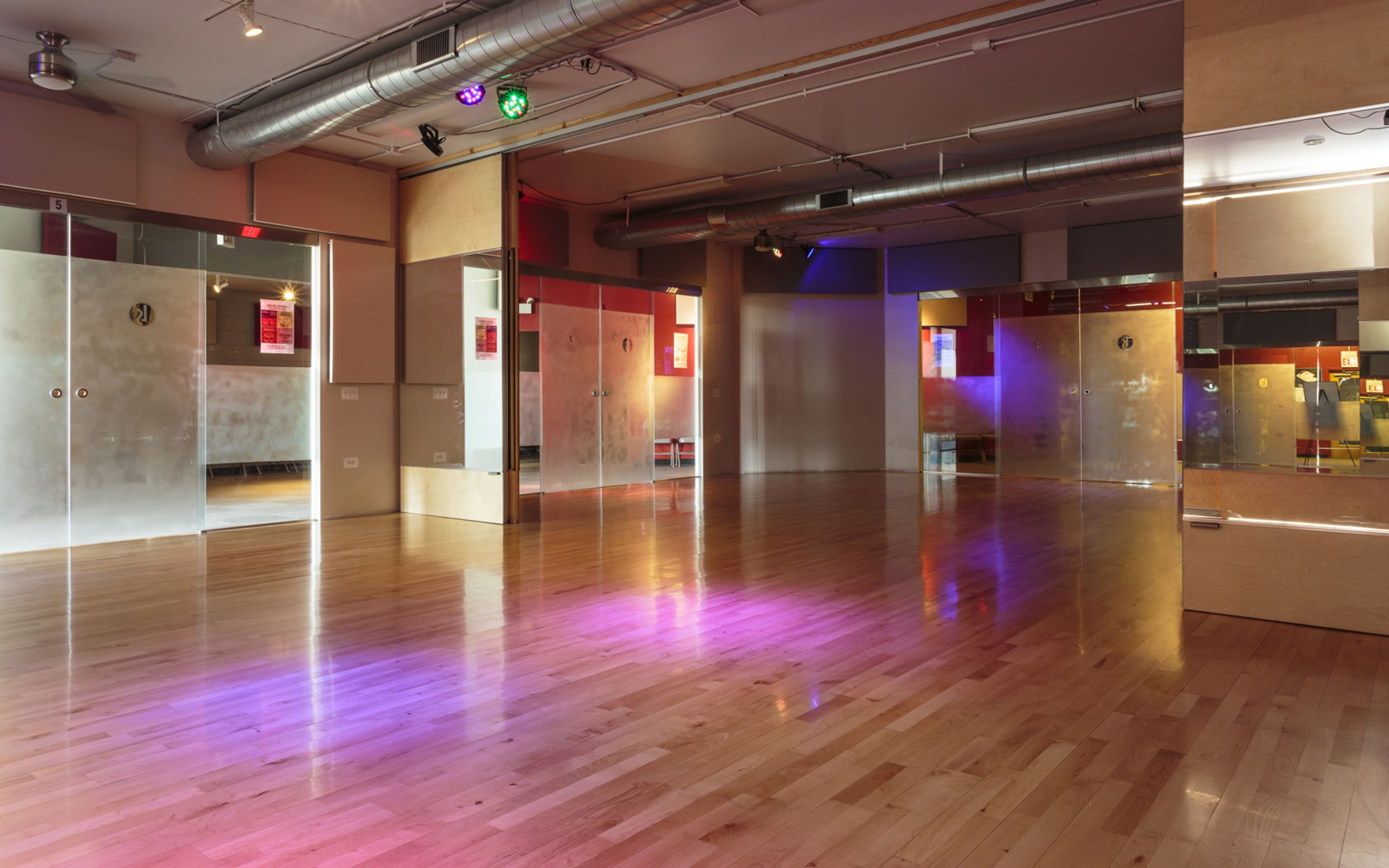 A spacious dance studio with wooden flooring, mirrored walls, and colorful lighting fixtures.