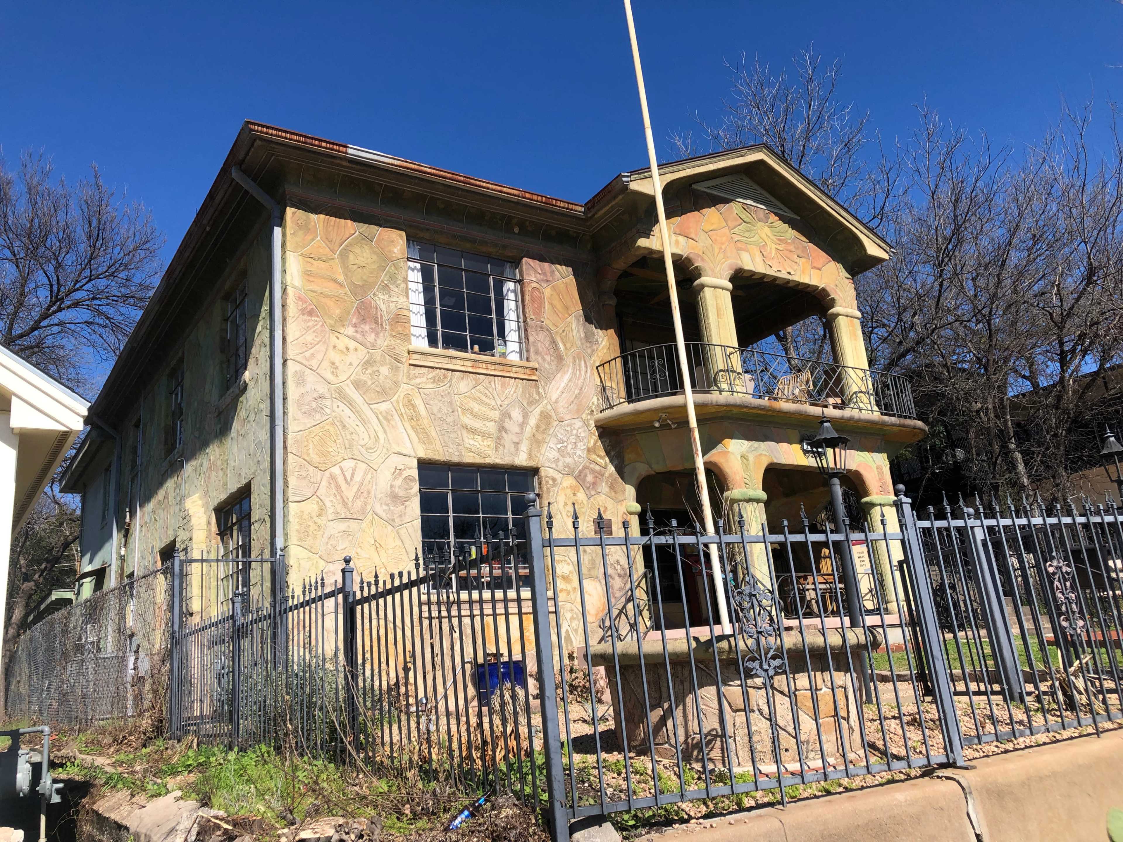 The image shows a two-story house with a stone-textured exterior and a balcony, surrounded by a metal fence and greenery.