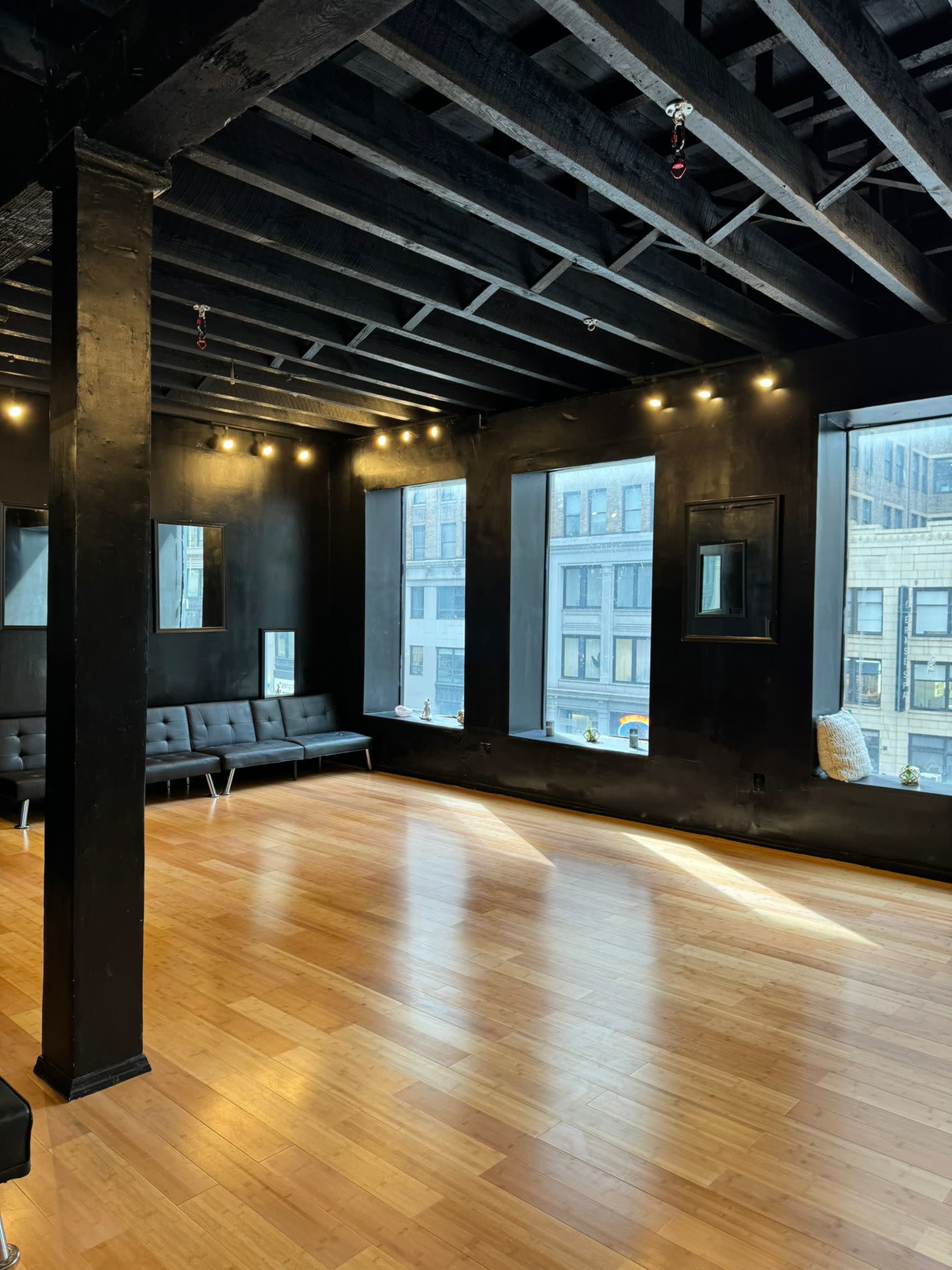 An empty room with a hardwood floor, black walls, and large windows allowing light to enter.