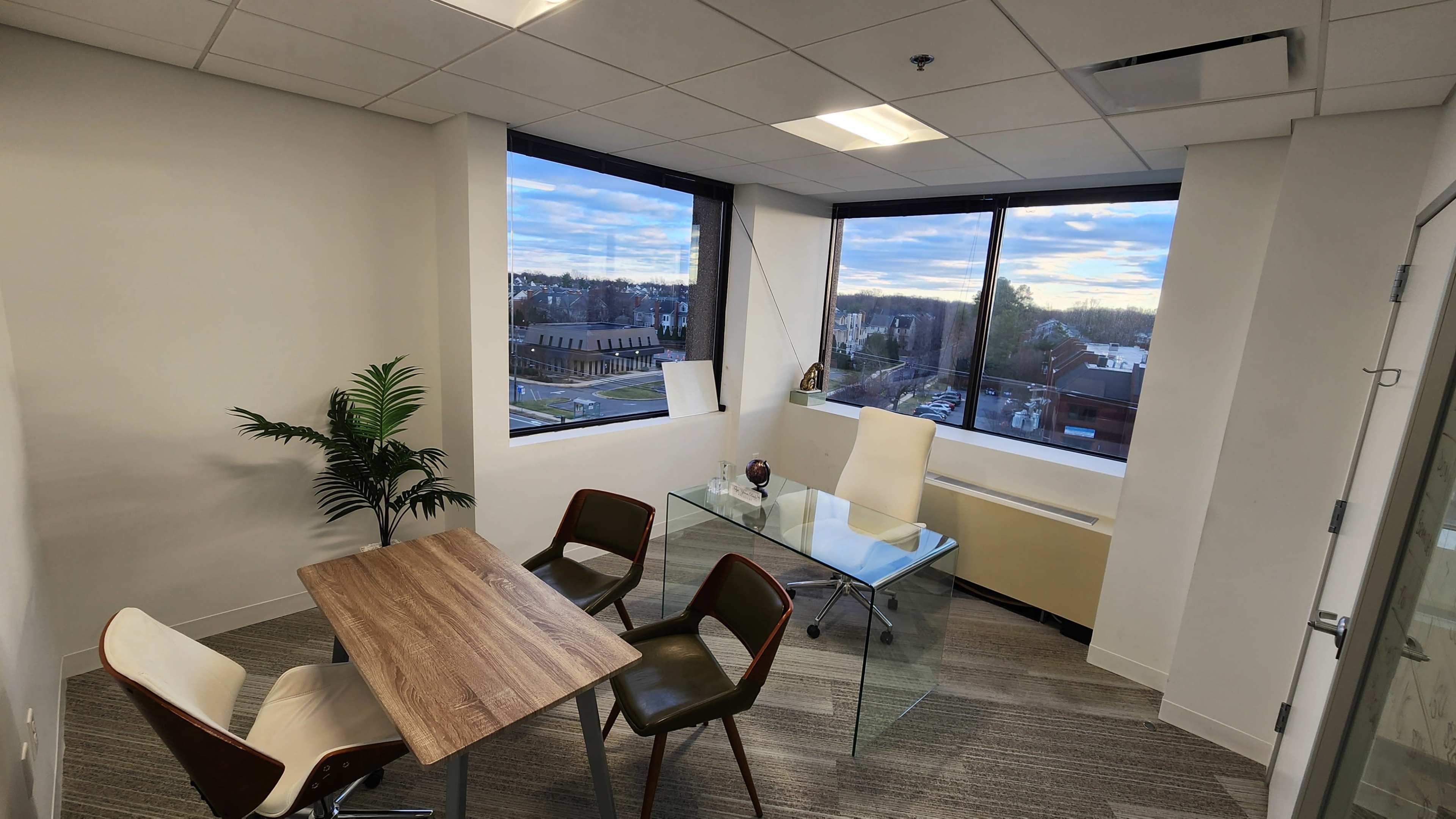 A small office space features a glass table and three chairs, with large windows overlooking a suburban area.