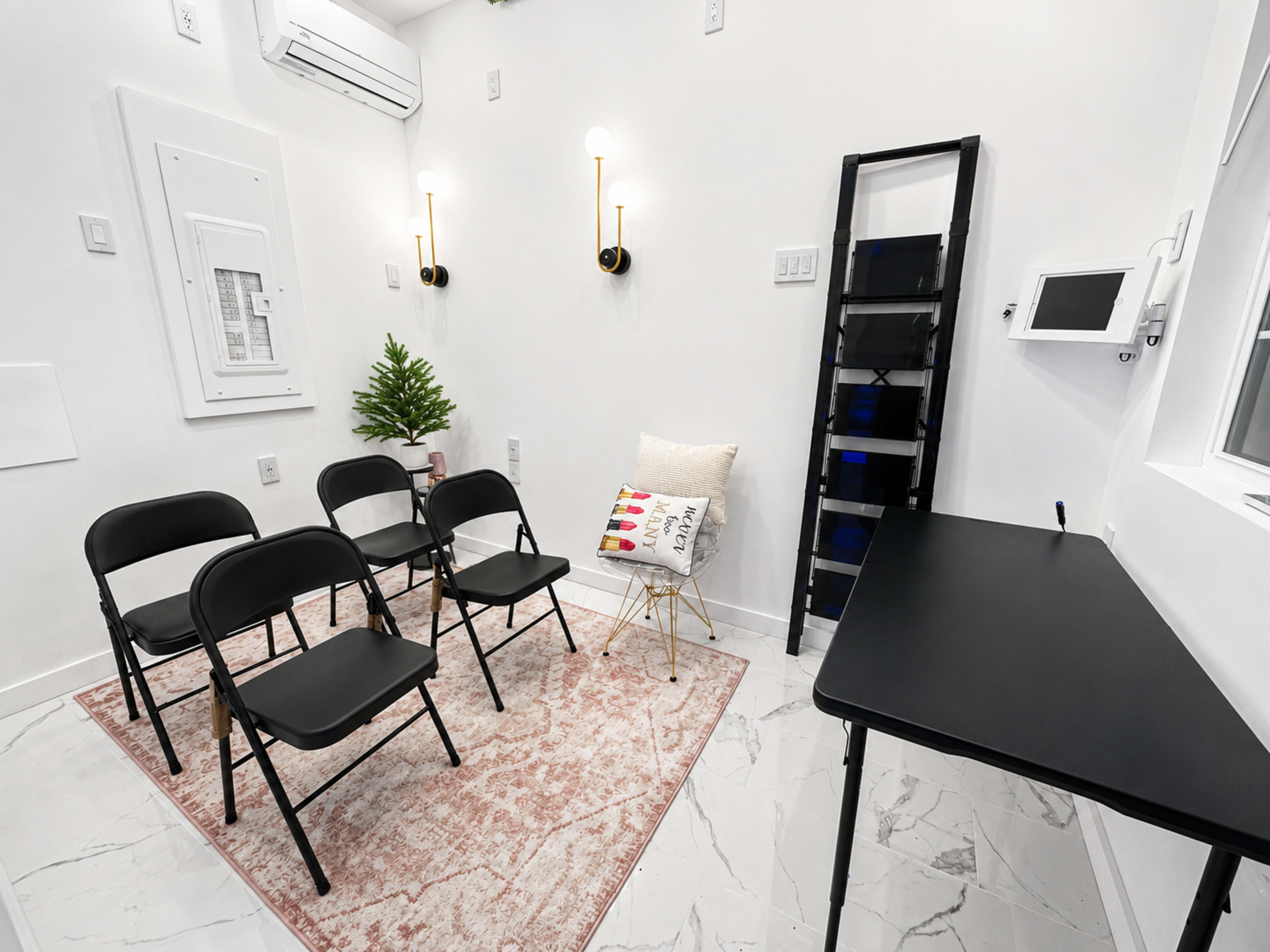The image shows a small, modern waiting room with four black folding chairs arranged in a row, a small plant, a decorative pillow on a chair, and a black table against a wall.