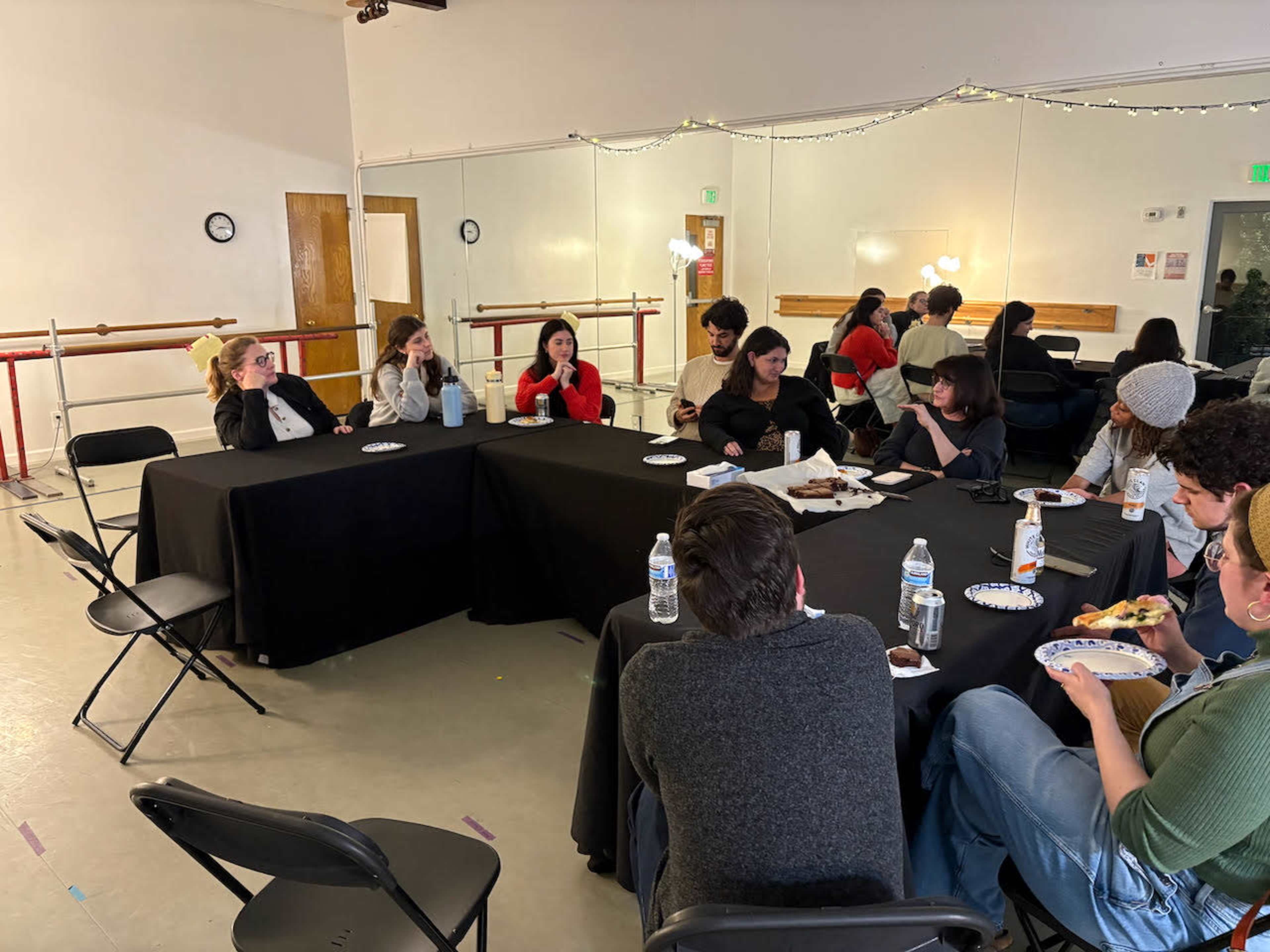 A group of people sits around black tables in a spacious room, engaged in conversation while sharing food and drinks.