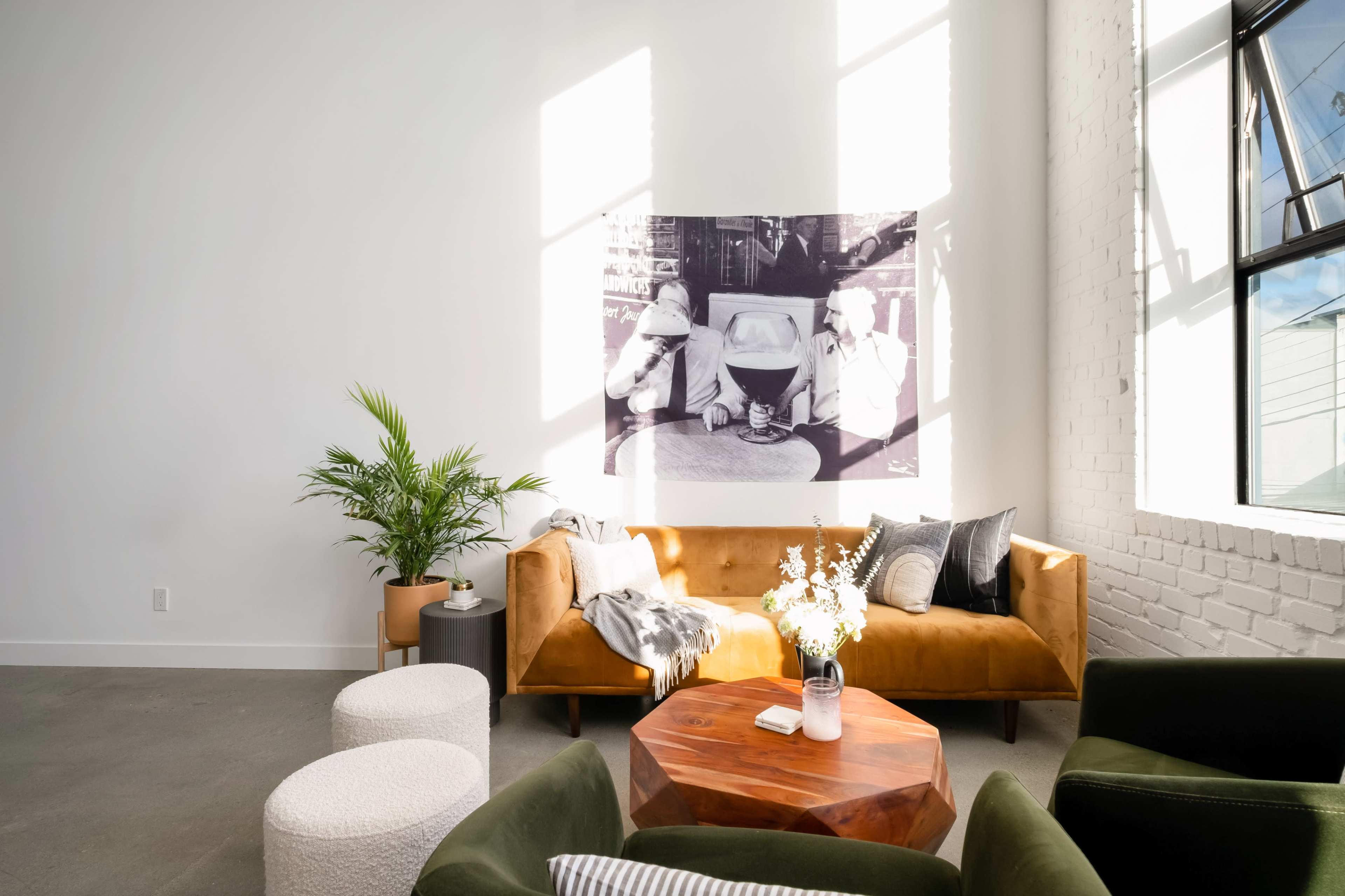 A spacious living room features a brown sofa, a round wooden coffee table, and a large black-and-white photograph on the wall, illuminated by natural light streaming through large windows.