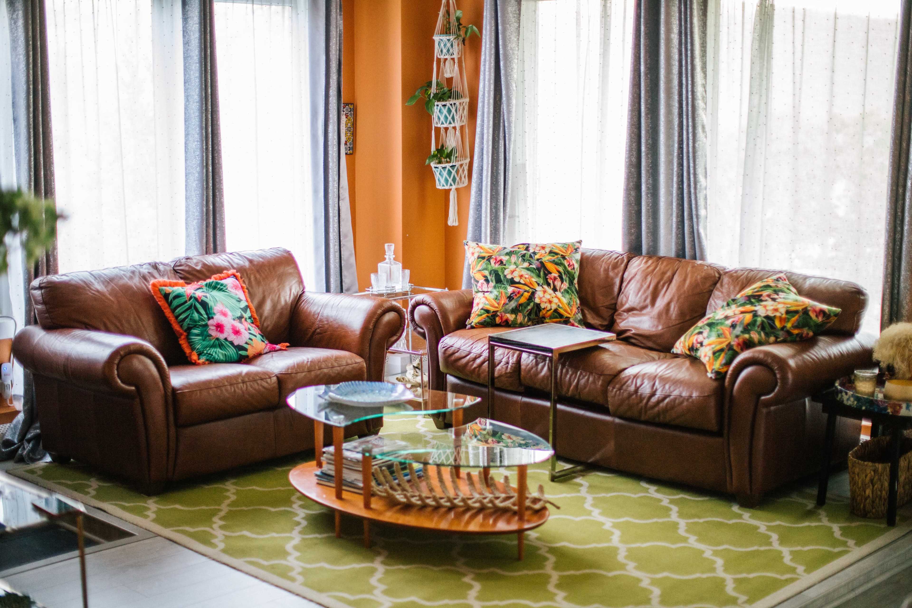 A cozy living room features two brown leather sofas adorned with colorful cushions, a glass coffee table, and large windows that let in natural light.