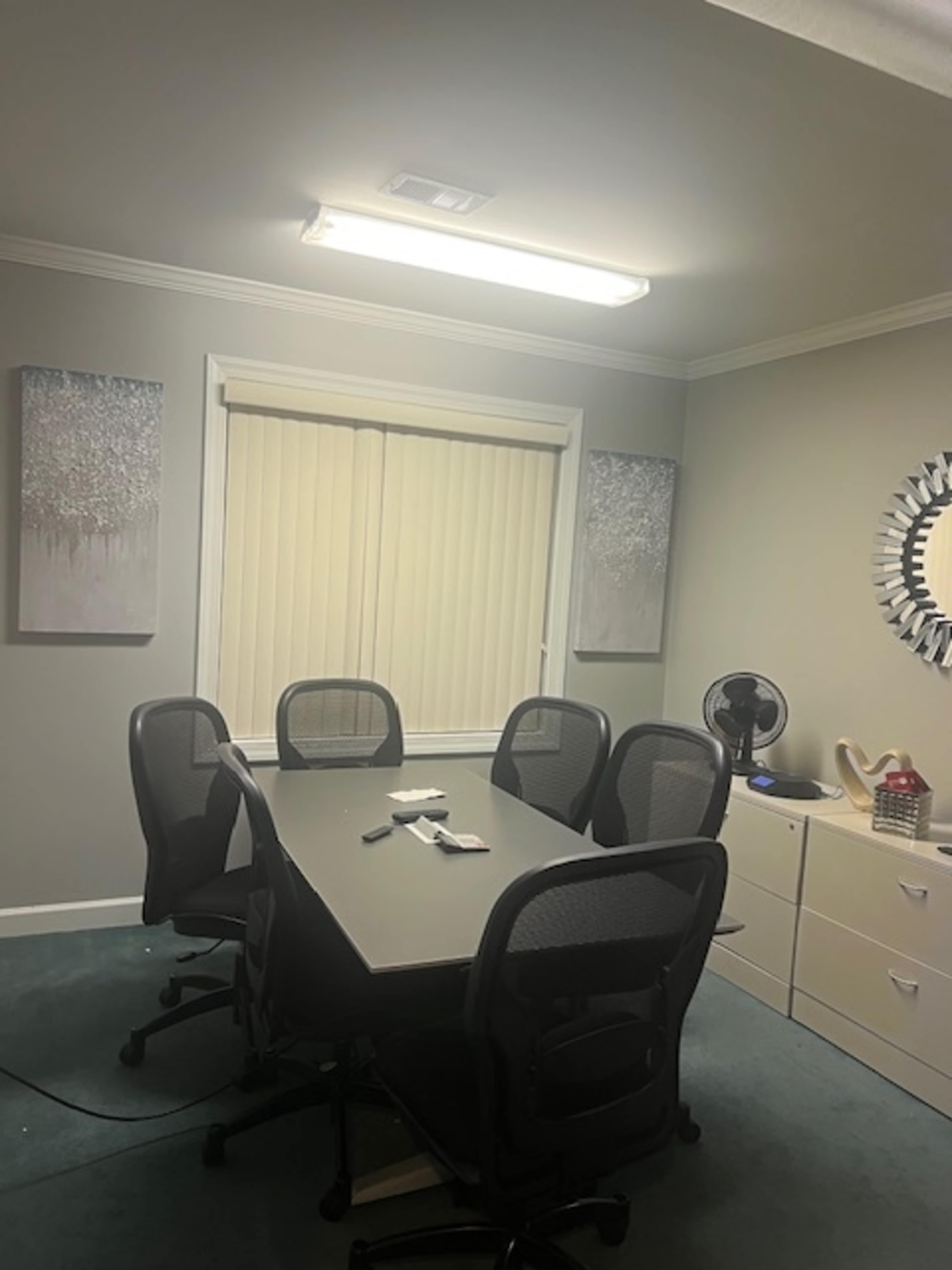 A conference room features a rectangular table surrounded by five black mesh chairs, with two framed artworks on the walls and a light fixture overhead.