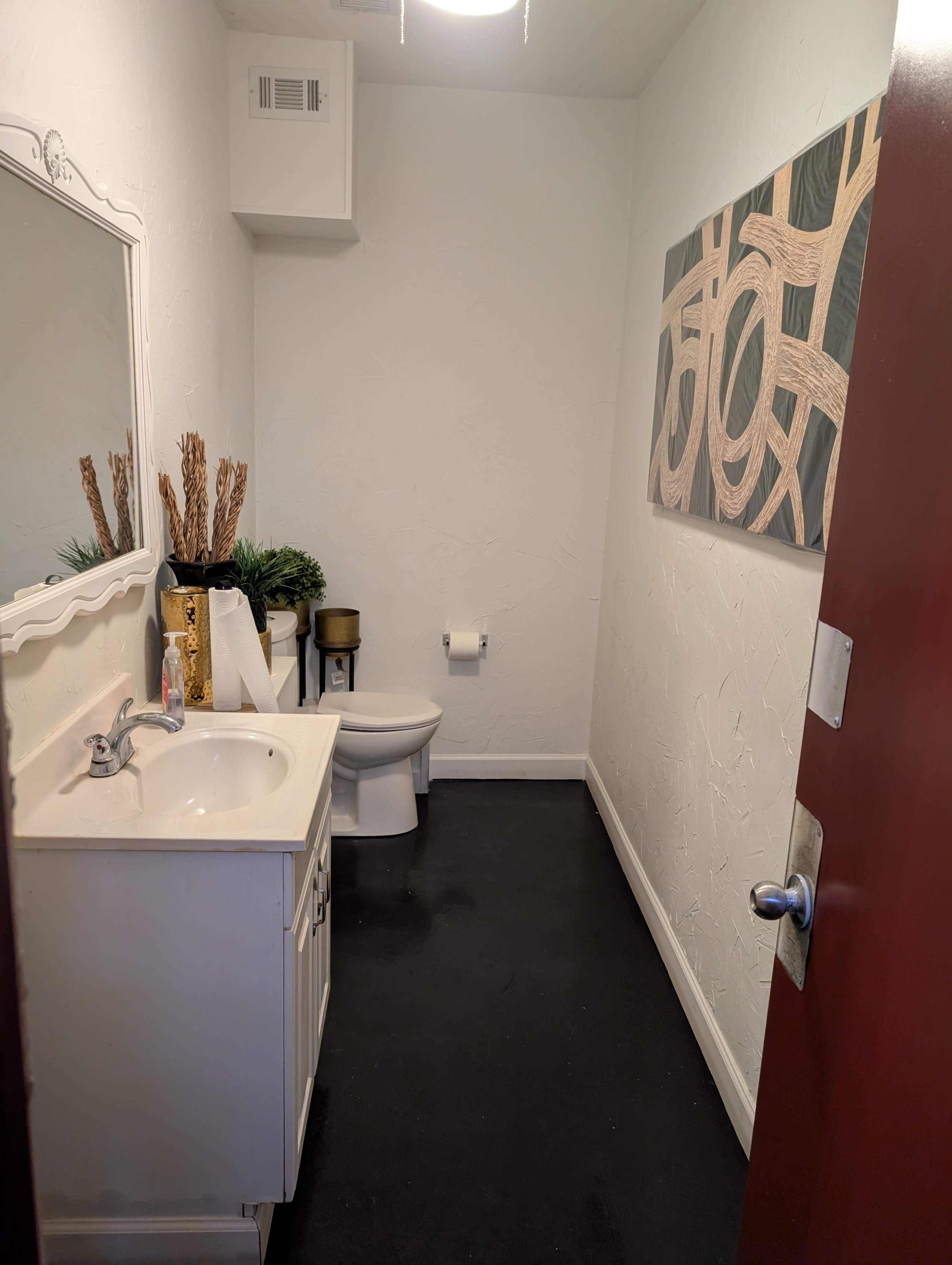 The image shows a small bathroom with a white sink and cabinet, a toilet, and a decorative wall art piece, all set against a light-colored wall and dark floor.
