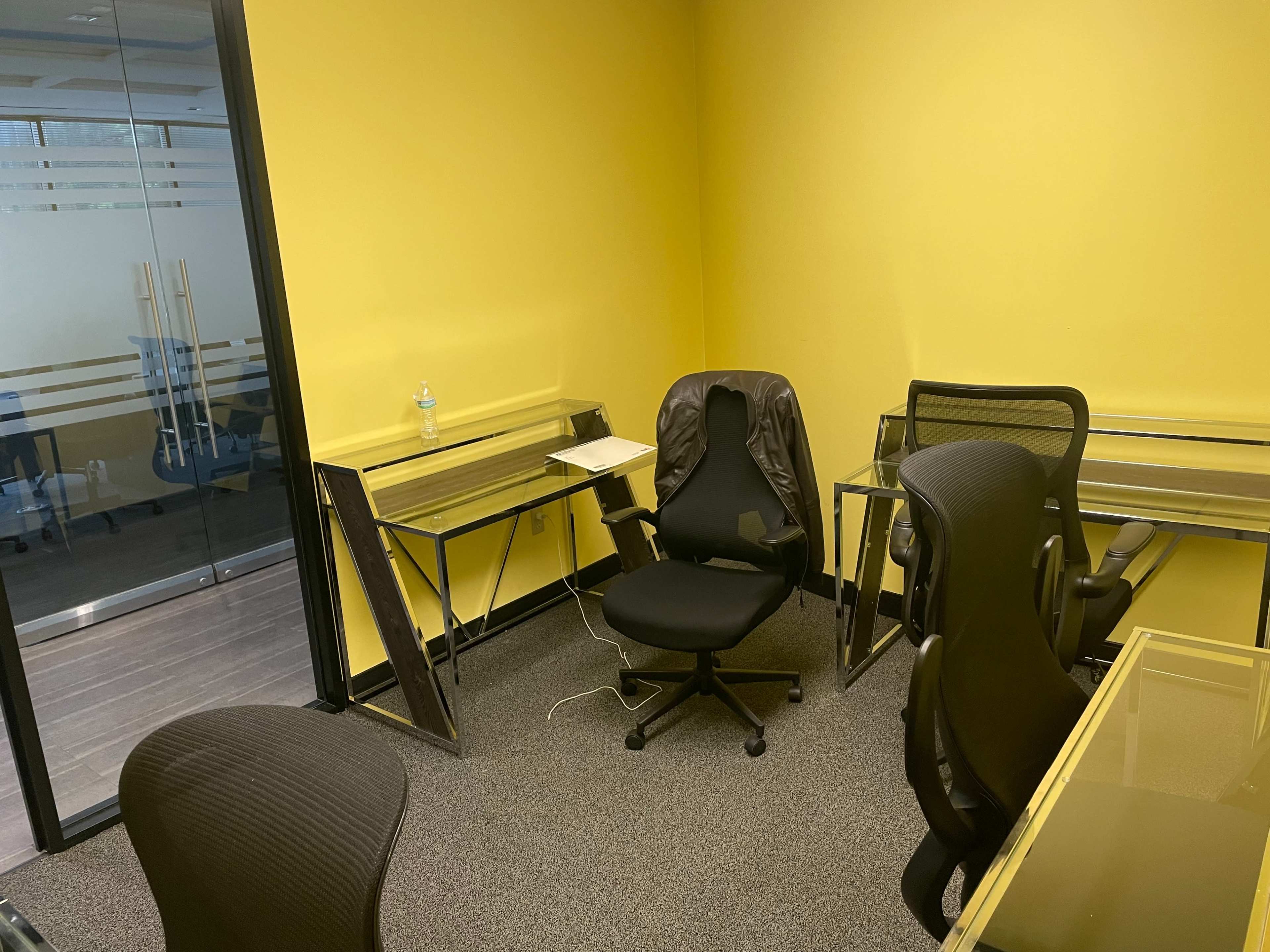 A small meeting room features two black office chairs and two glass-top desks against bright yellow walls.