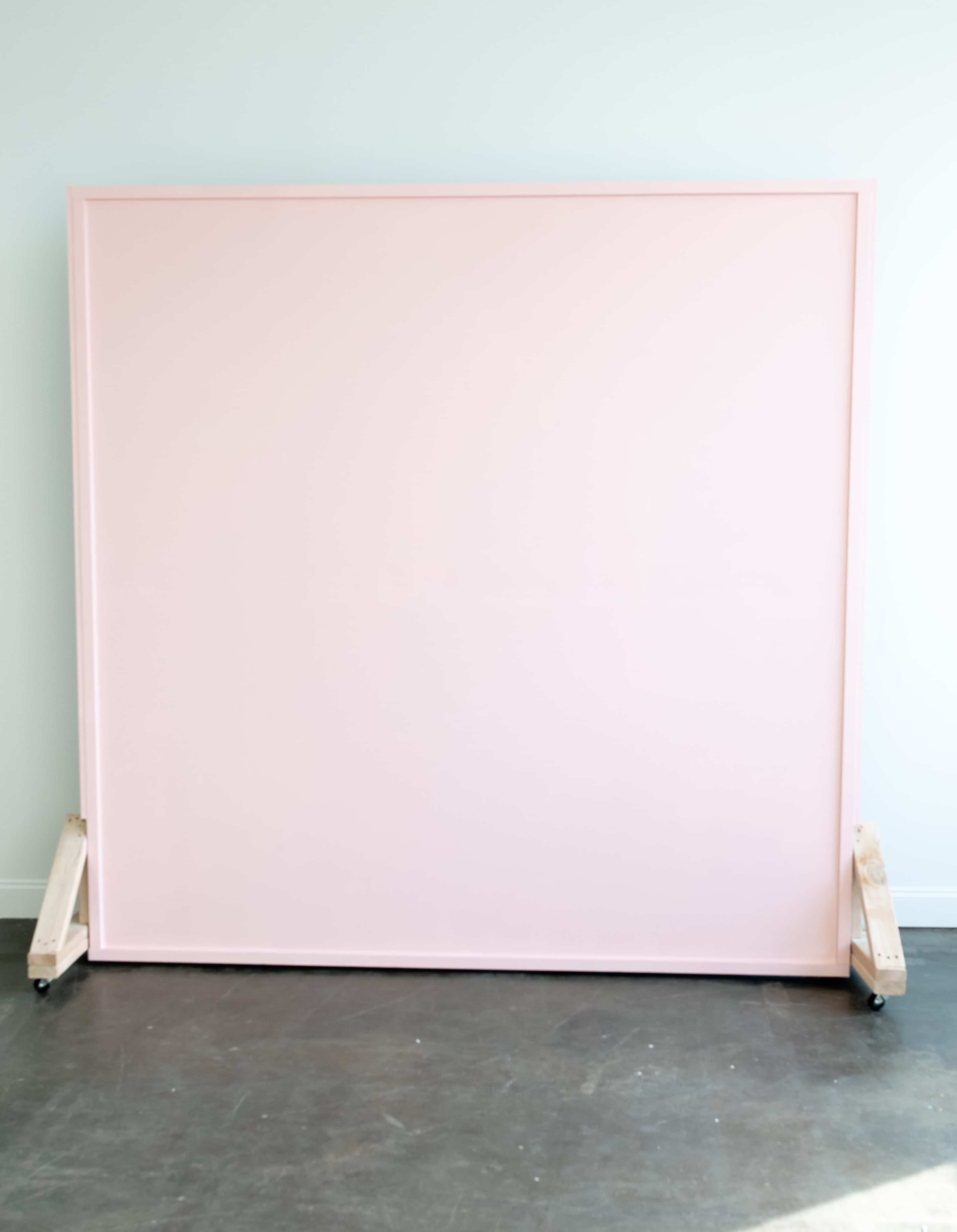 The image shows a large, pink canvas mounted on wooden supports with wheels, positioned against a light-colored wall.