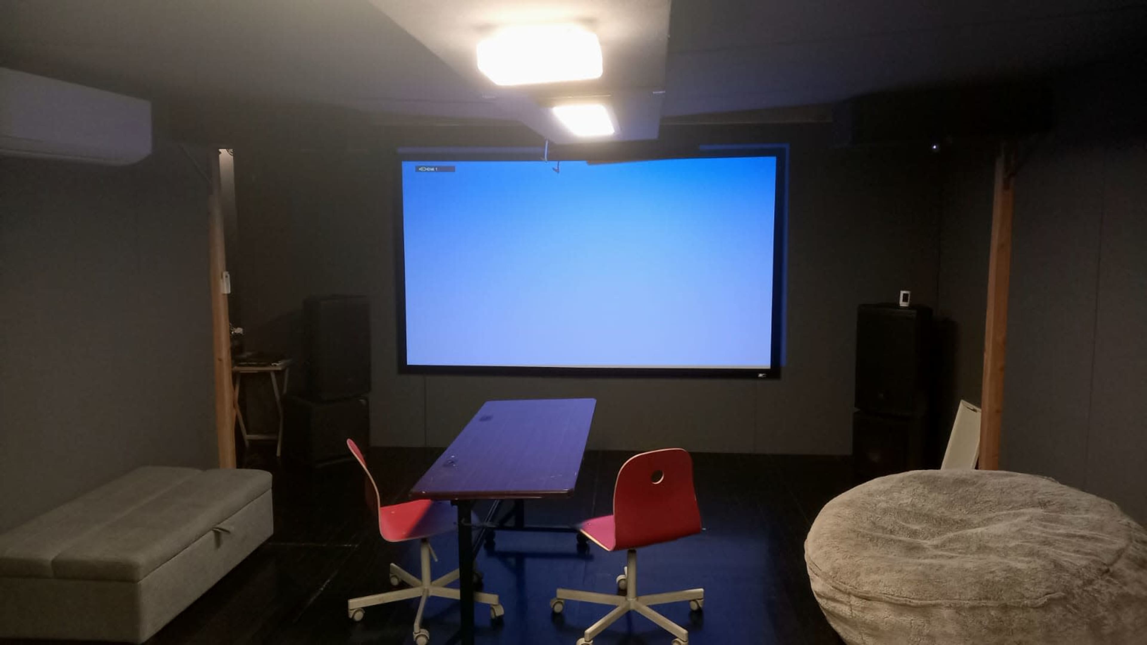 A dimly lit room with a large blank screen on one wall, a table in the center, and two red chairs placed around it.