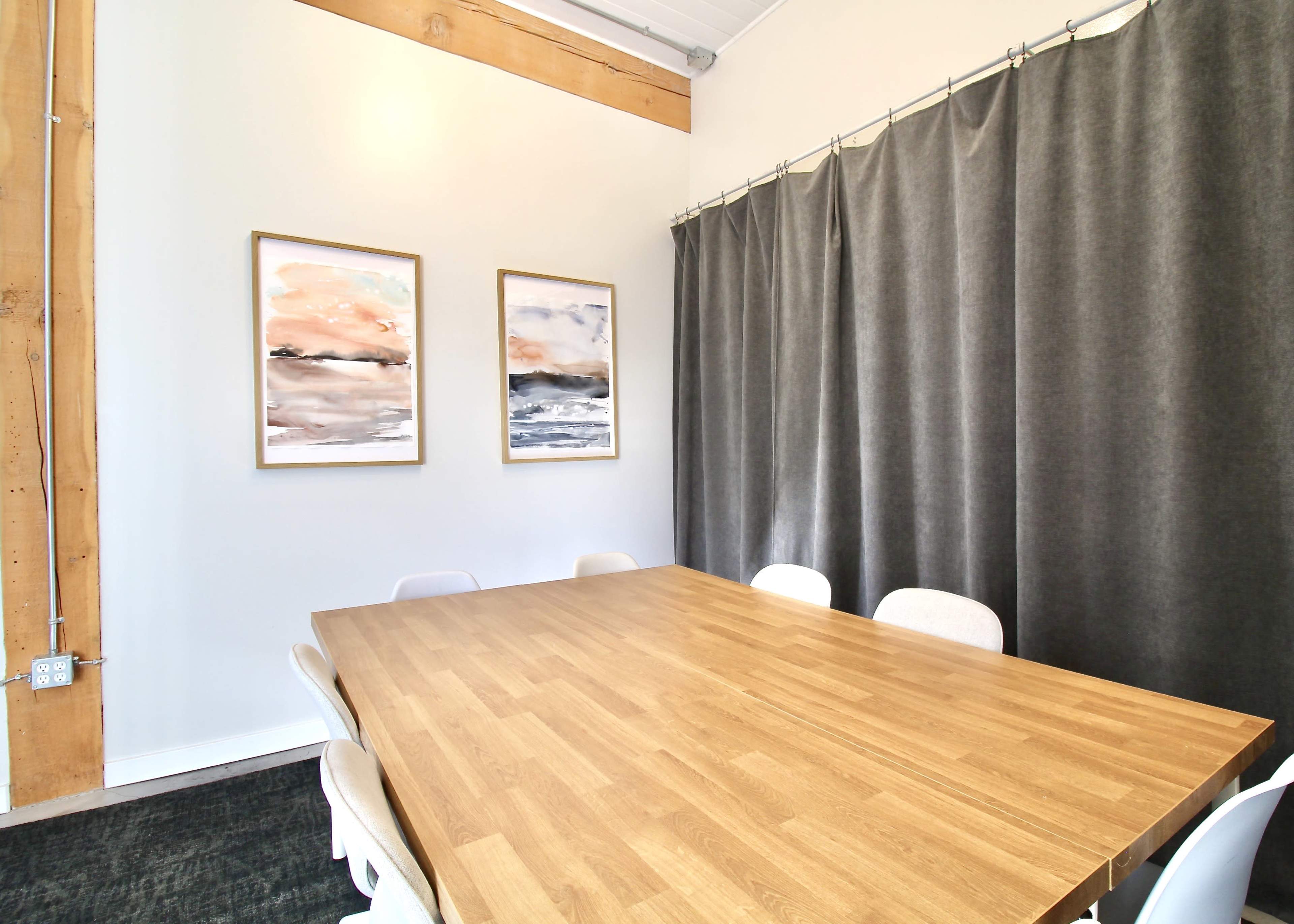 A wooden conference table with several chairs is positioned in a meeting room featuring grey curtains and two framed paintings on the wall.