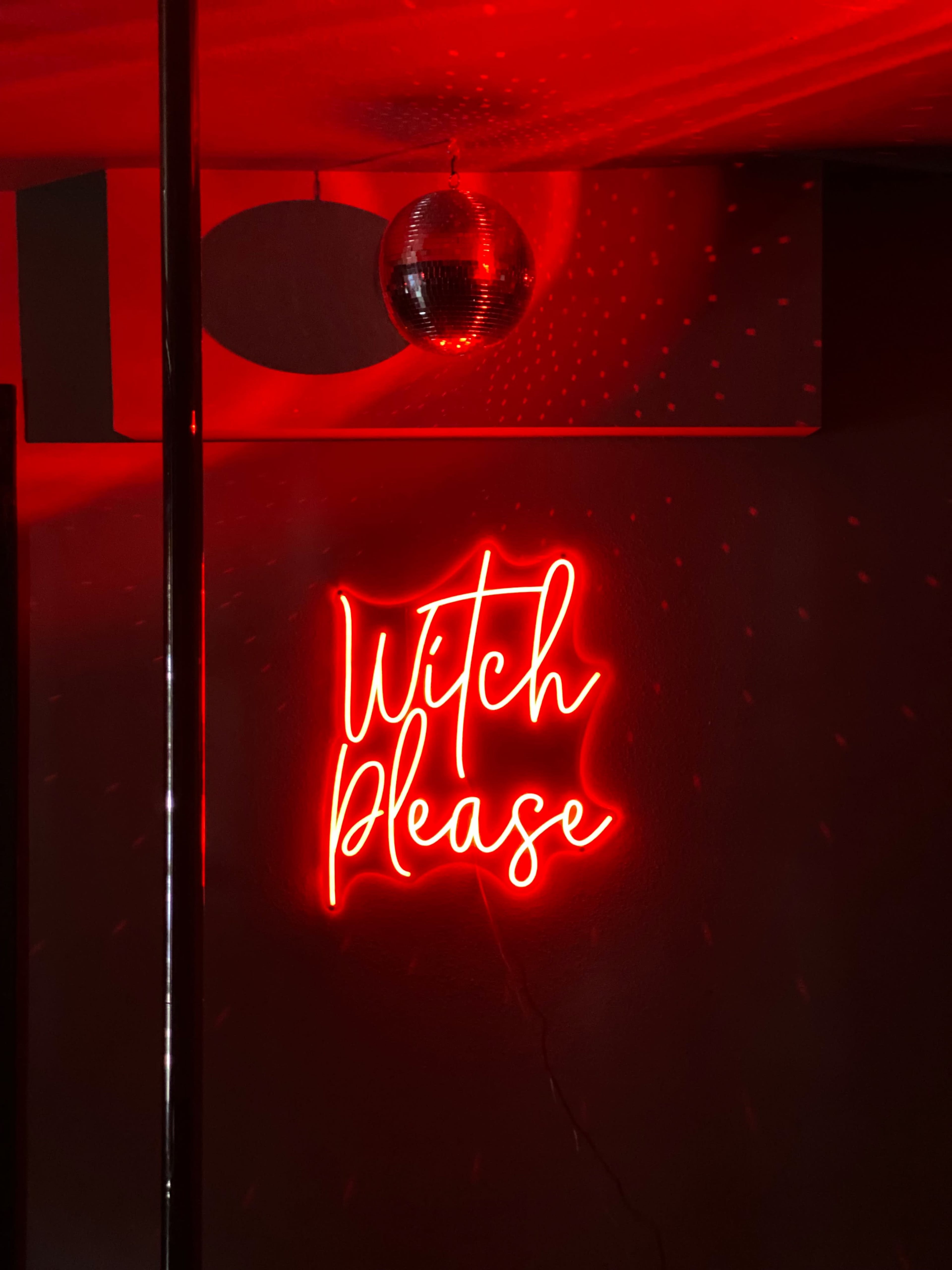 A red neon sign reading "Witch Please" is displayed on a dark wall with a disco ball reflecting light.