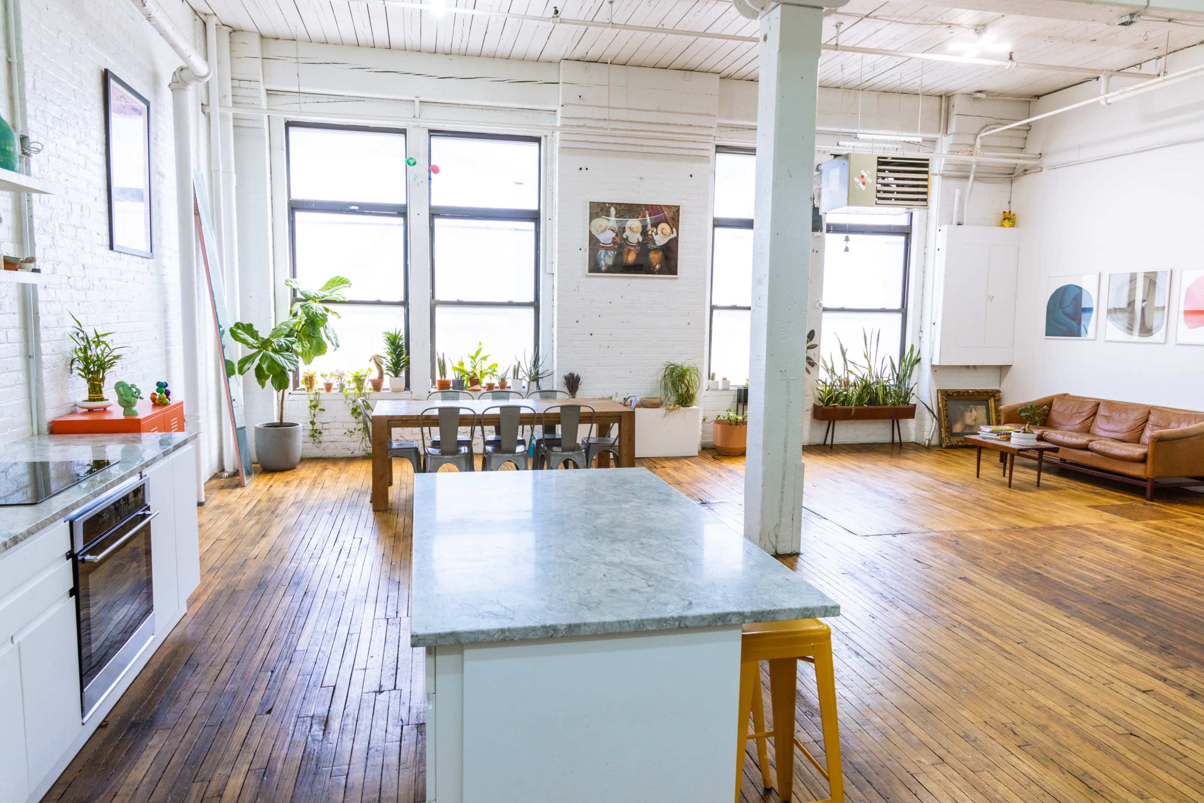 A spacious kitchen and living area with wood flooring, large windows, plants, a dining table, and a leather sofa.