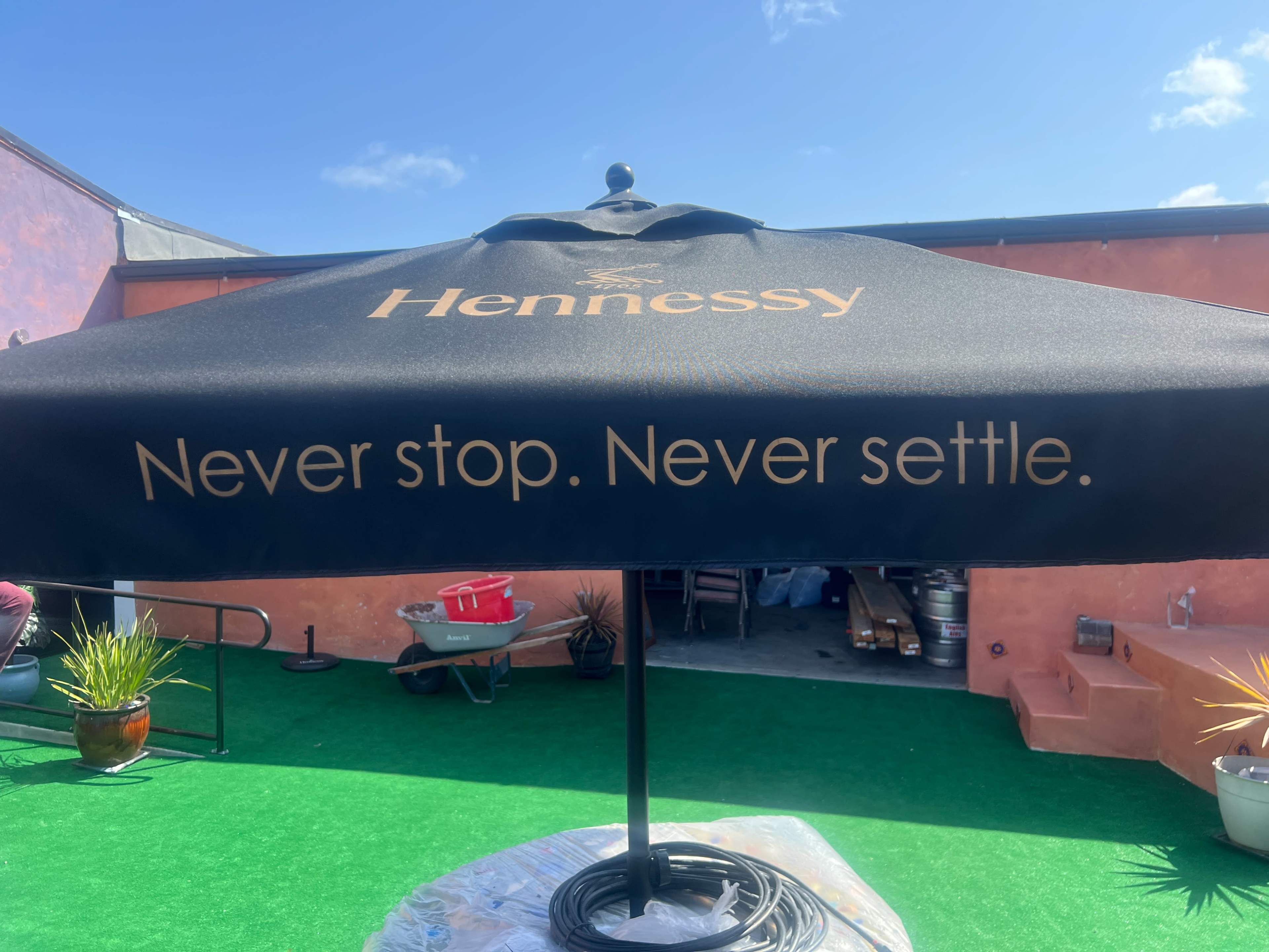 The image shows a black umbrella featuring the Hennessy logo and the phrase "Never stop. Never settle."