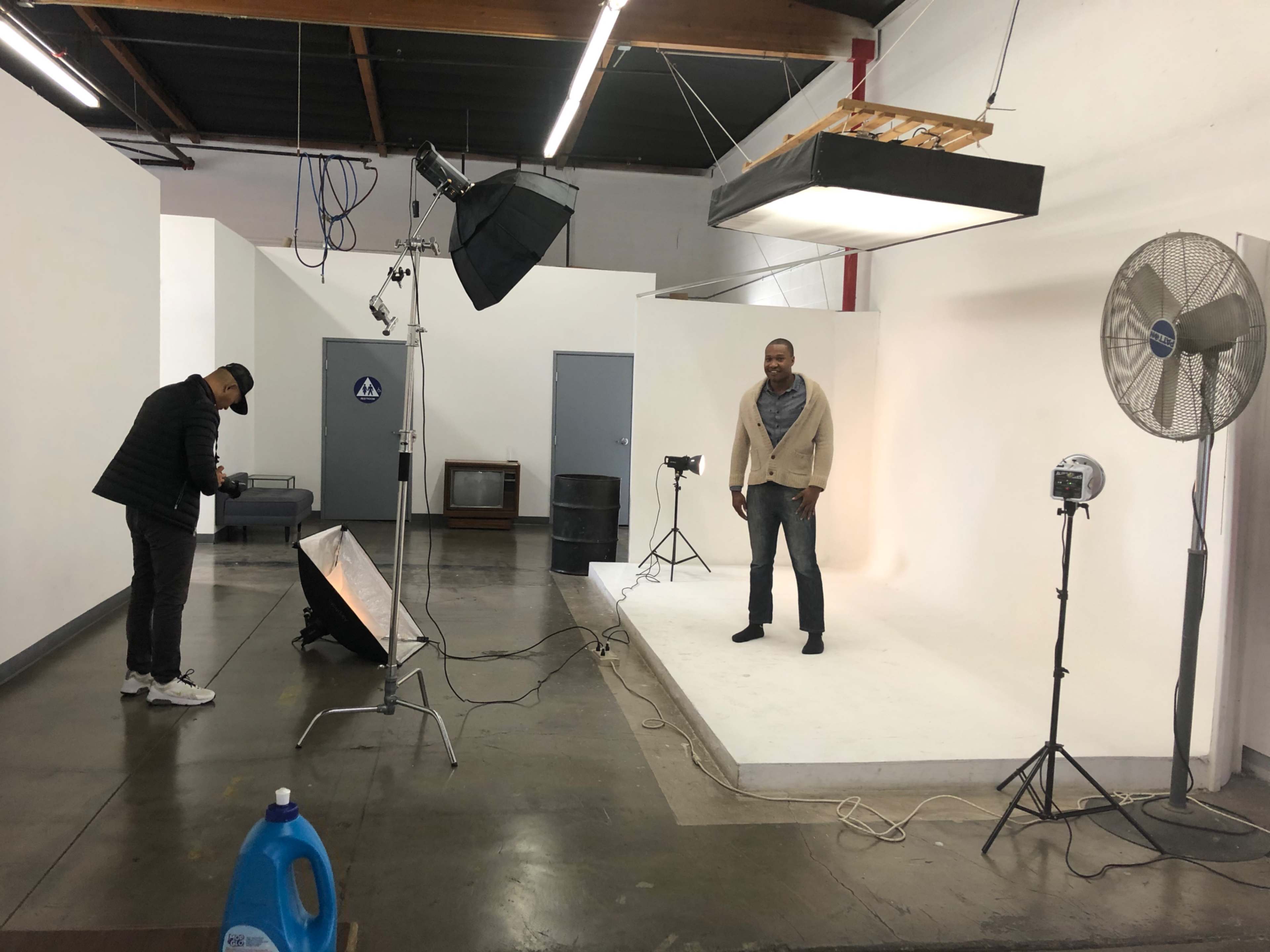 A man is standing on a white backdrop in a photography studio, while another person sets up equipment nearby.