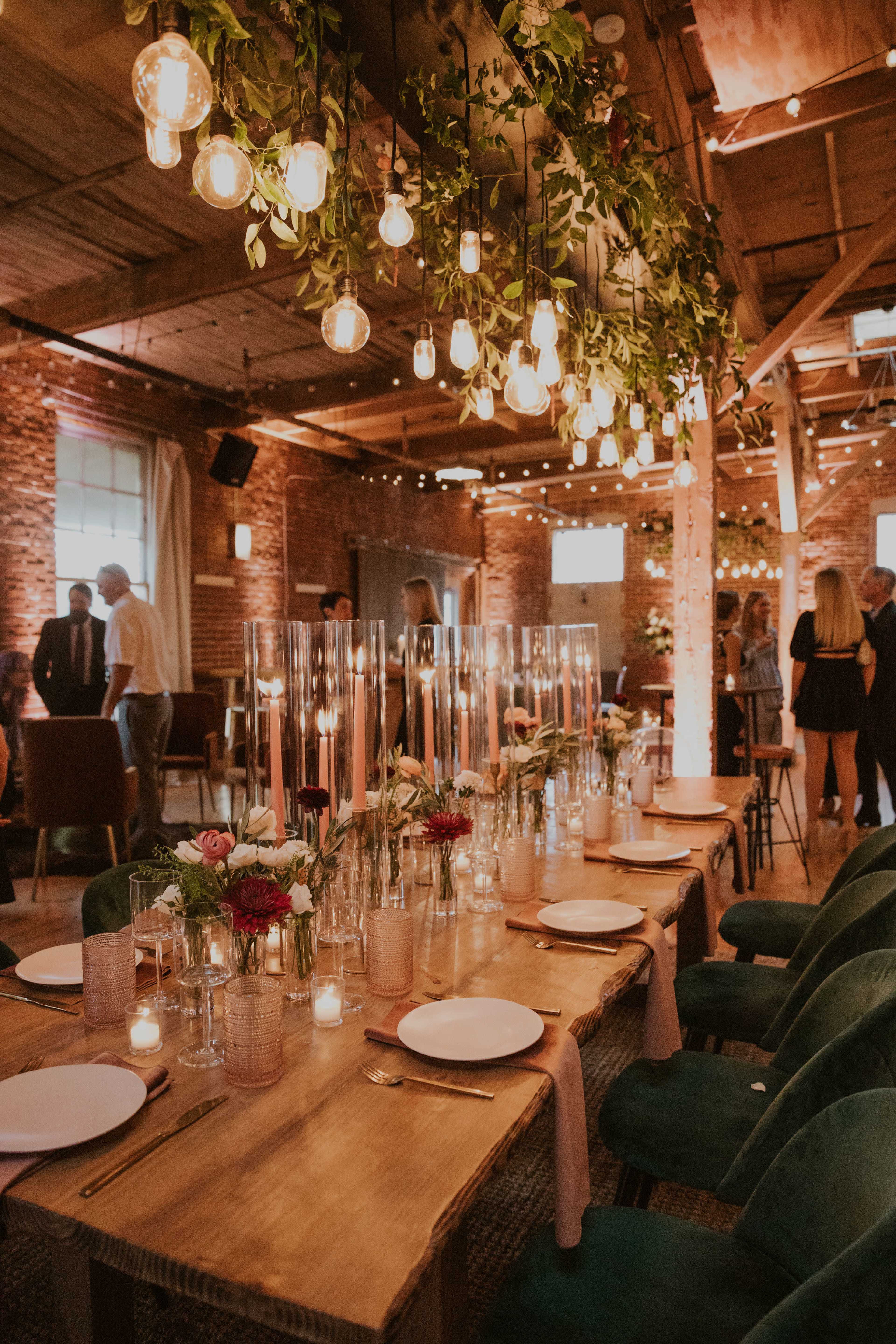 A long wooden dining table is set with candles, plates, and glass vases of flowers under hanging string lights in a rustic event space.