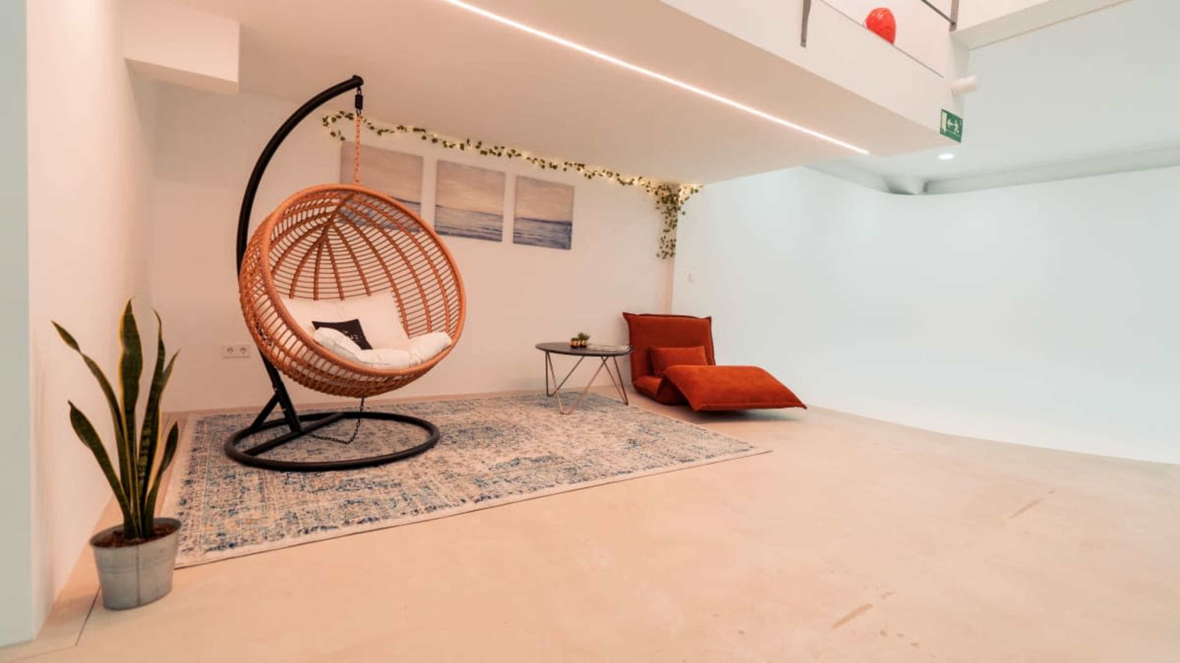 The image shows a modern, minimalistic interior space featuring a woven hanging chair, a small table, and a red lounge cushion on a beige floor.