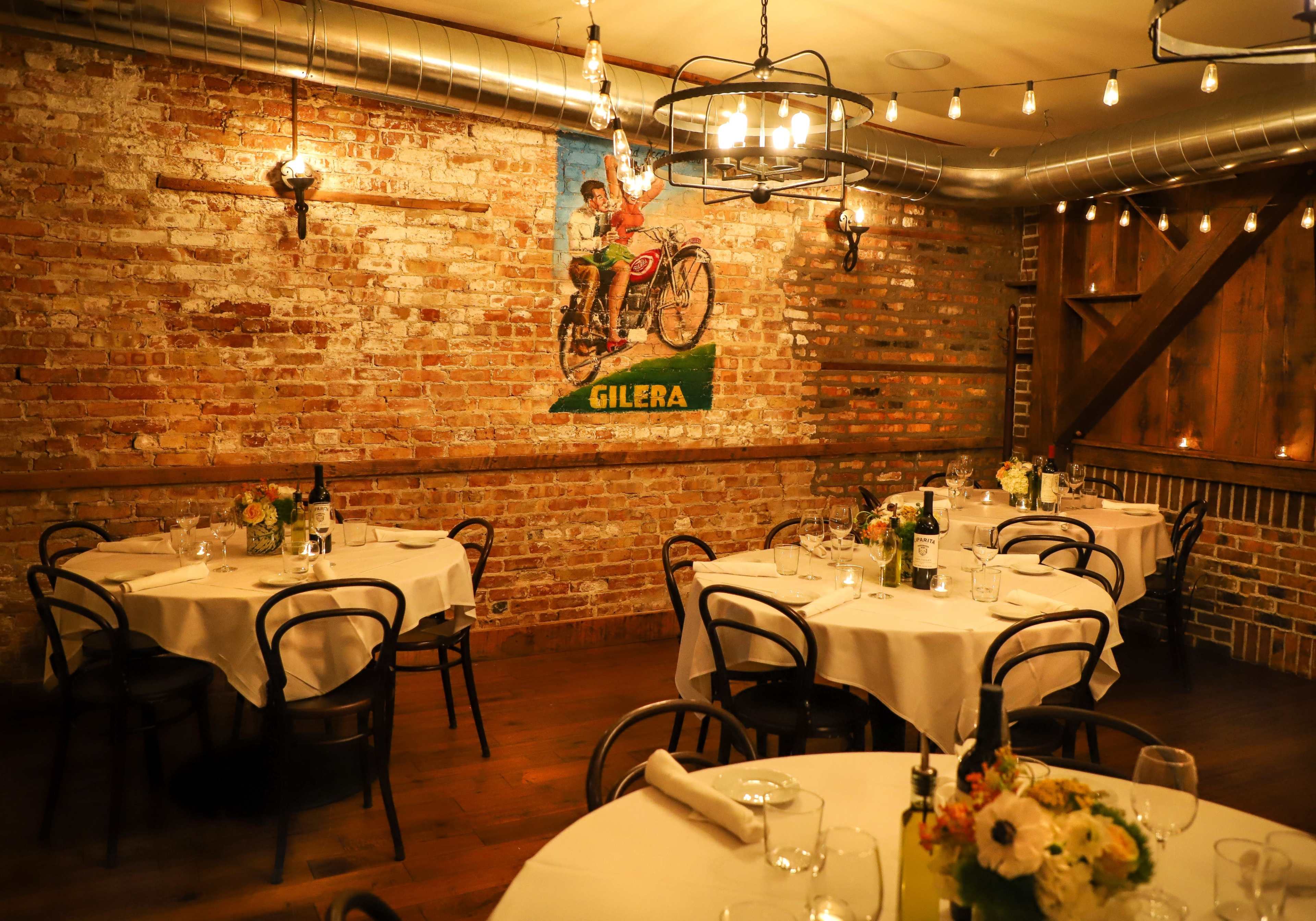 The image shows a cozy restaurant interior with wooden tables, black chairs, exposed brick walls, and a vintage Gilera motorcycle advertisement.