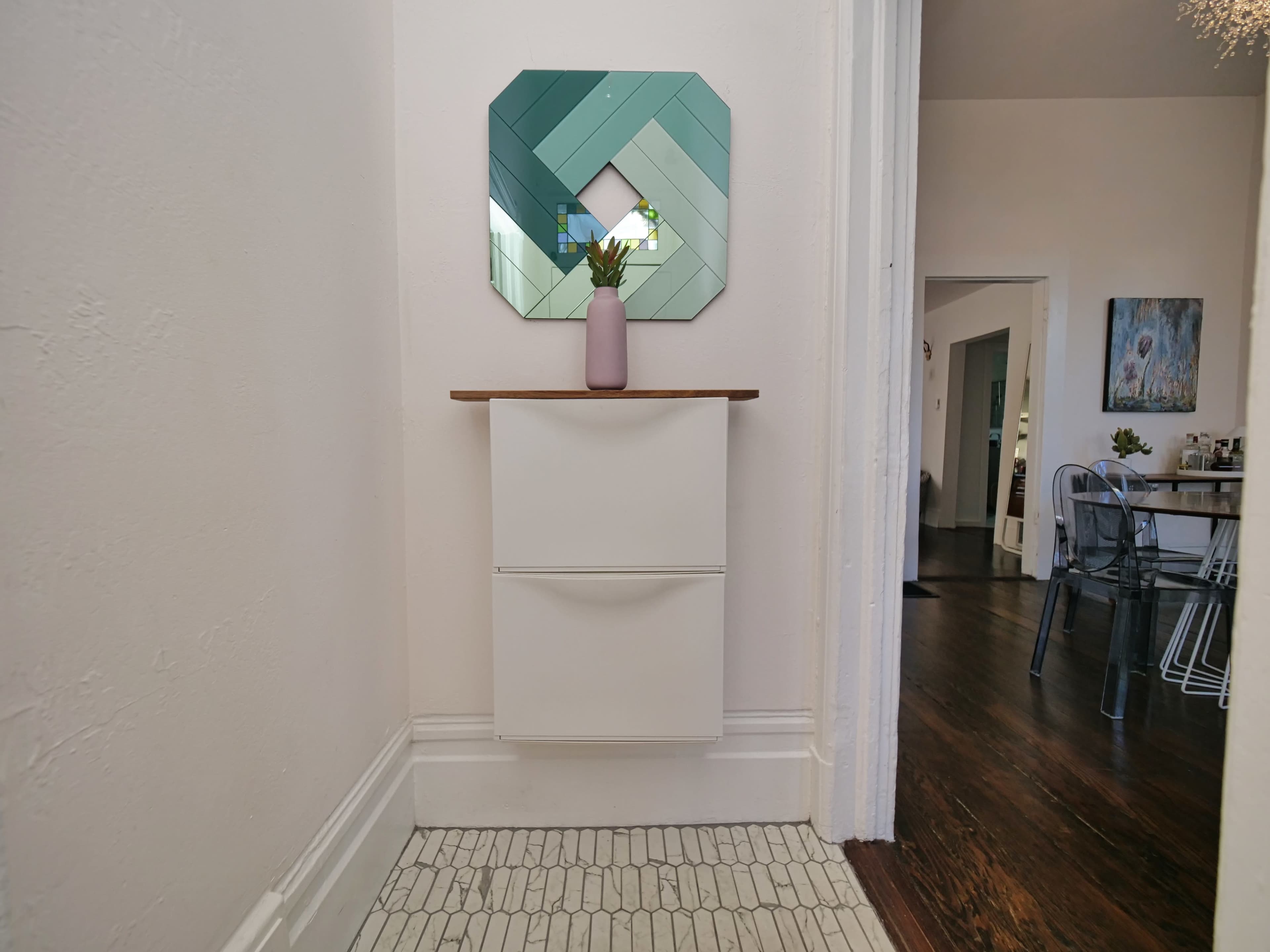 A narrow entryway features a white cabinet with two drawers beneath a geometric mirror and a vase with a plant on top.