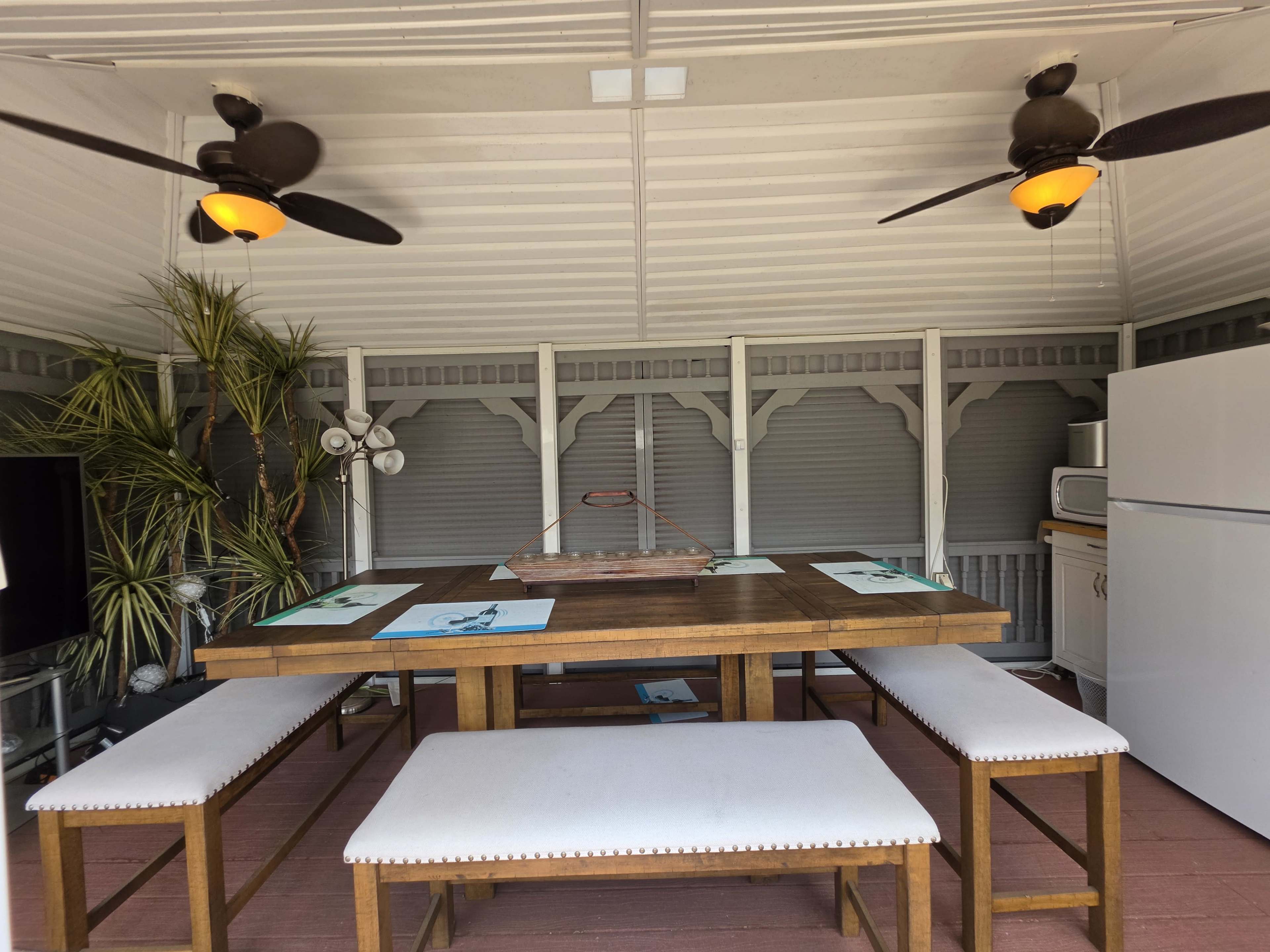 The image shows a covered outdoor dining area with a large wooden table, benches, ceiling fans, and a refrigerator in a shaded environment.
