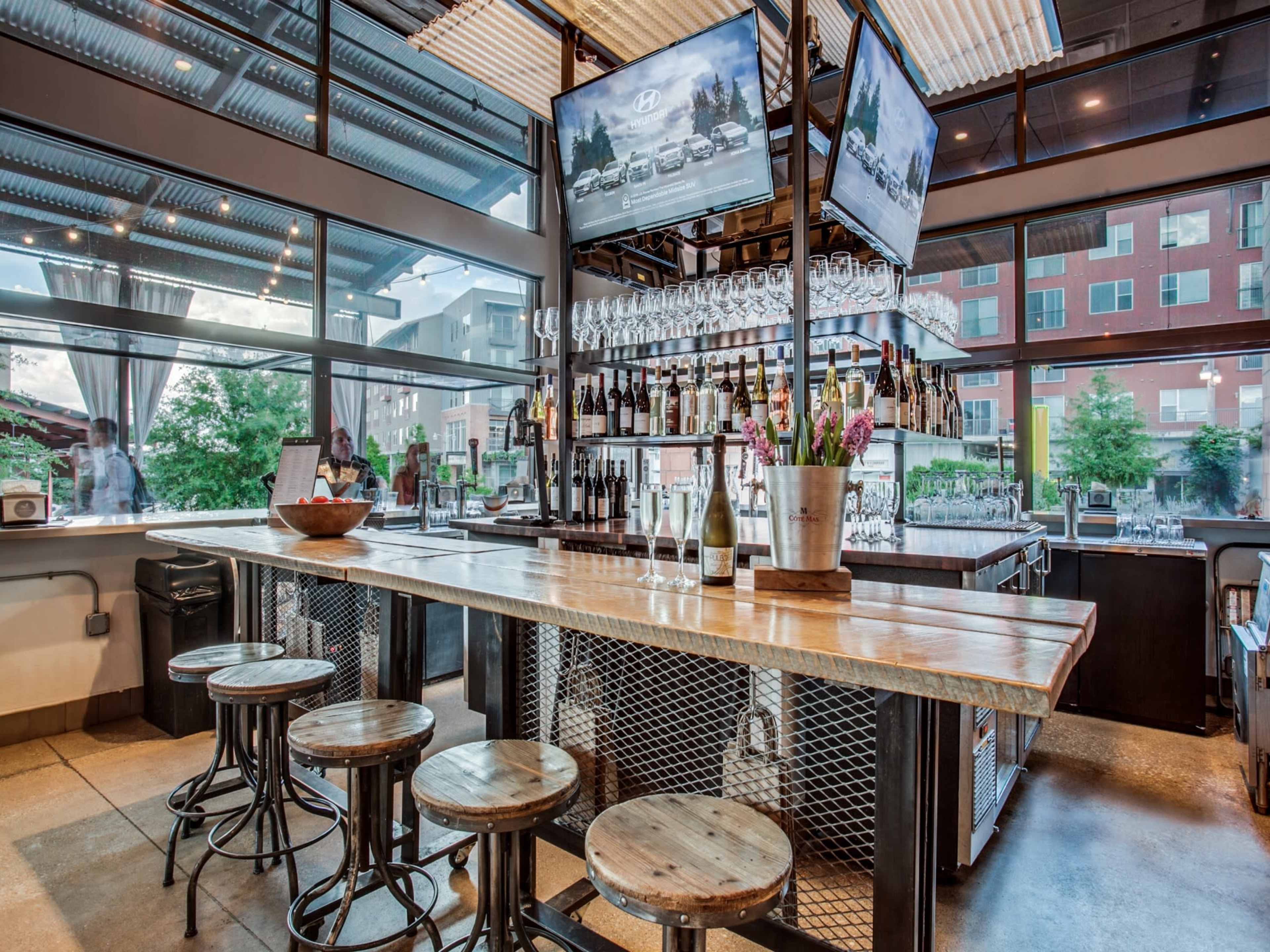 A modern bar with a rustic wooden counter, metal stools, and multiple televisions displaying content, surrounded by large windows.