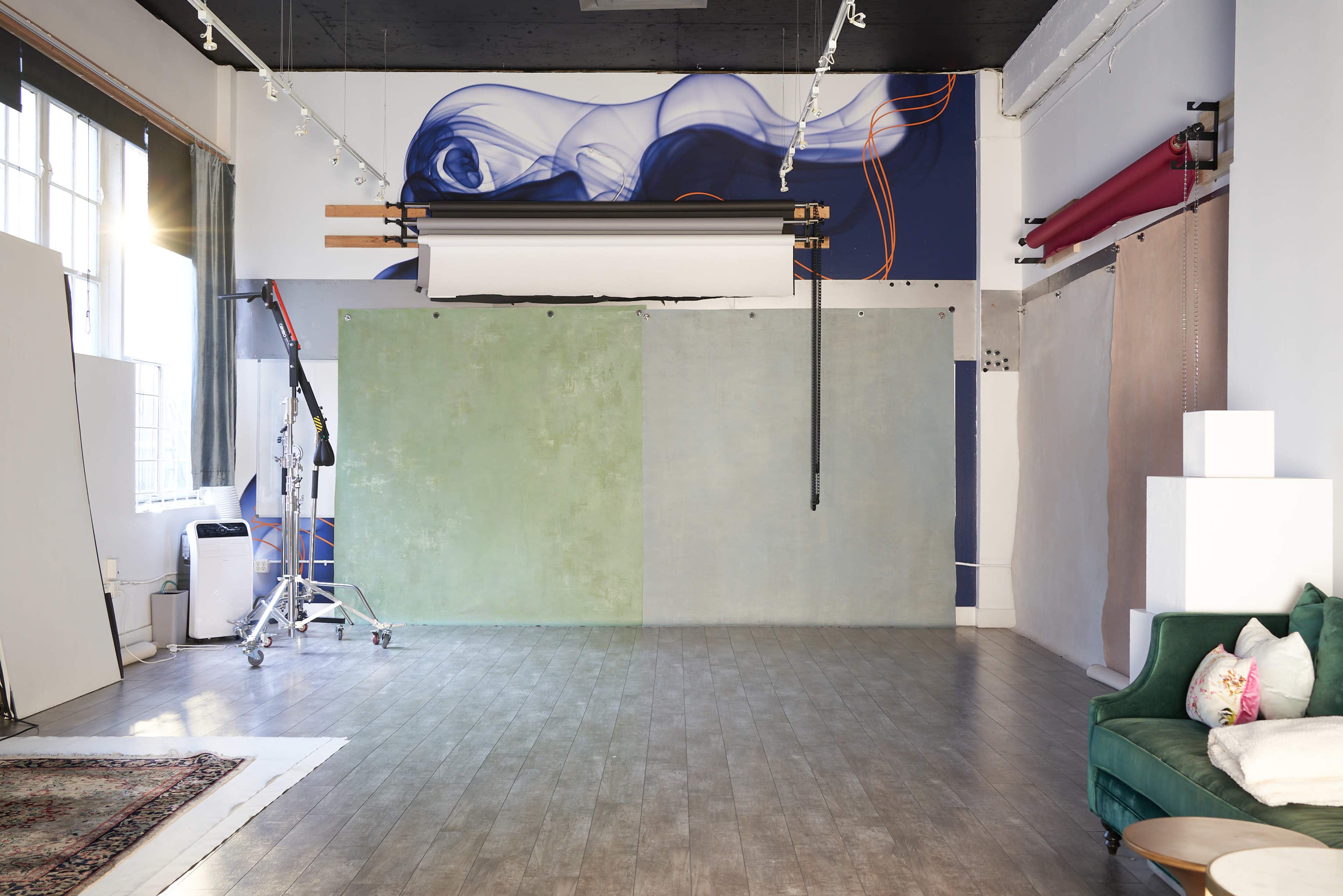 The image shows a spacious photo studio featuring a green and blue backdrop, a large lighting setup, and a green couch in a well-lit area.