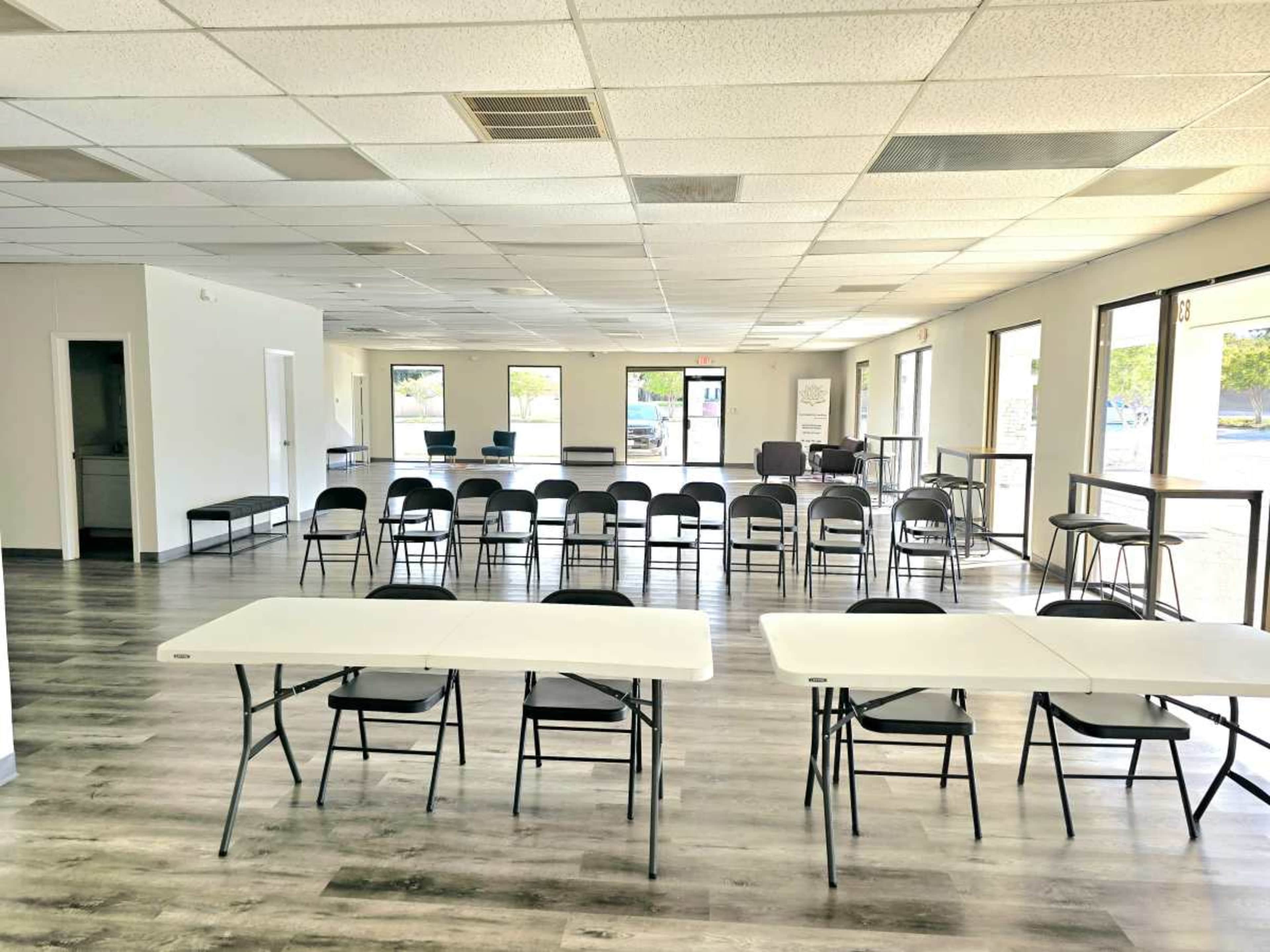 The image shows a spacious, empty room with rows of black chairs and several white folding tables arranged in the center.
