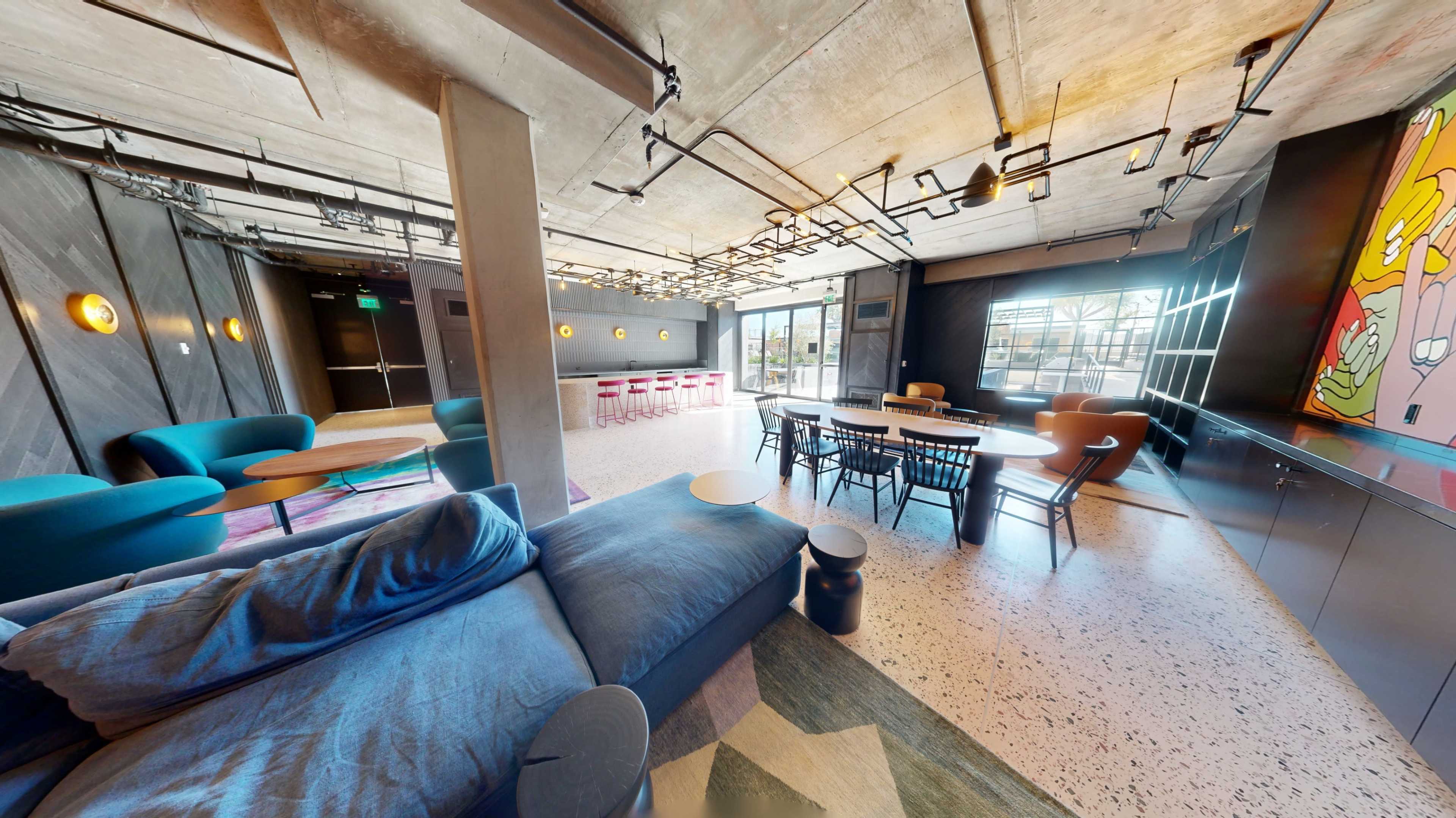 The image shows a modern open-space lounge area with comfortable seating, a dining table, and large windows allowing natural light.