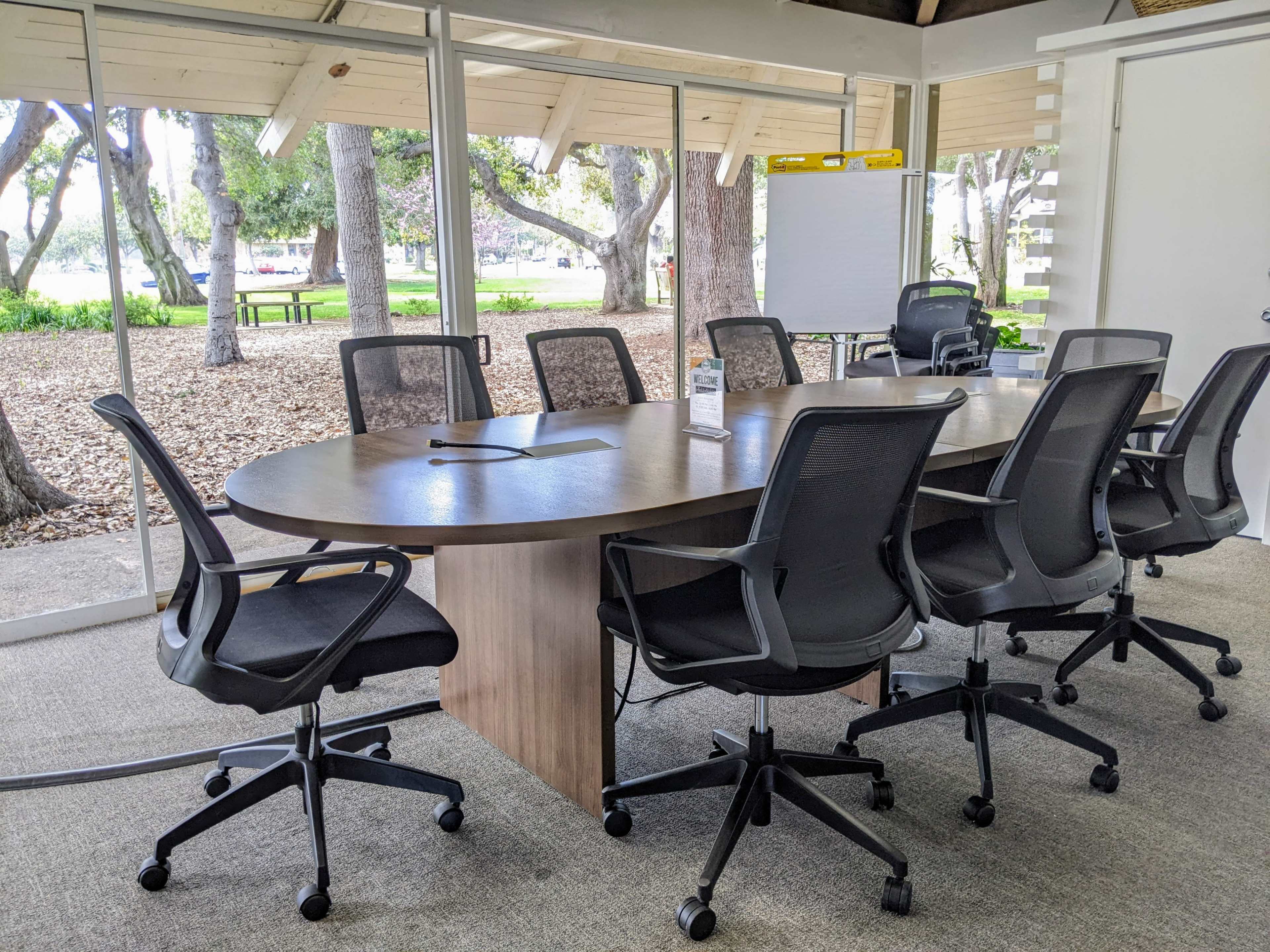 A conference room features a large oval table surrounded by six black rolling chairs, with large windows showing a view of trees and an outdoor area.