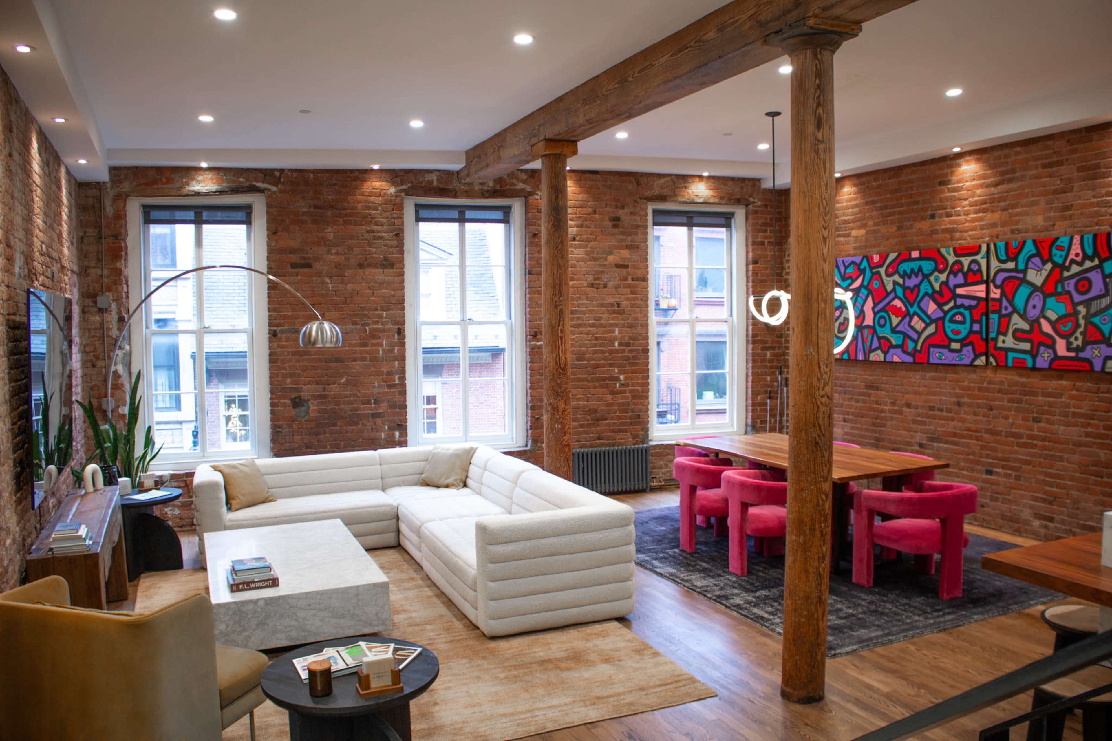 The image shows a modern living area featuring a large sectional sofa, a wooden dining table with pink chairs, exposed brick walls, and wooden pillars.