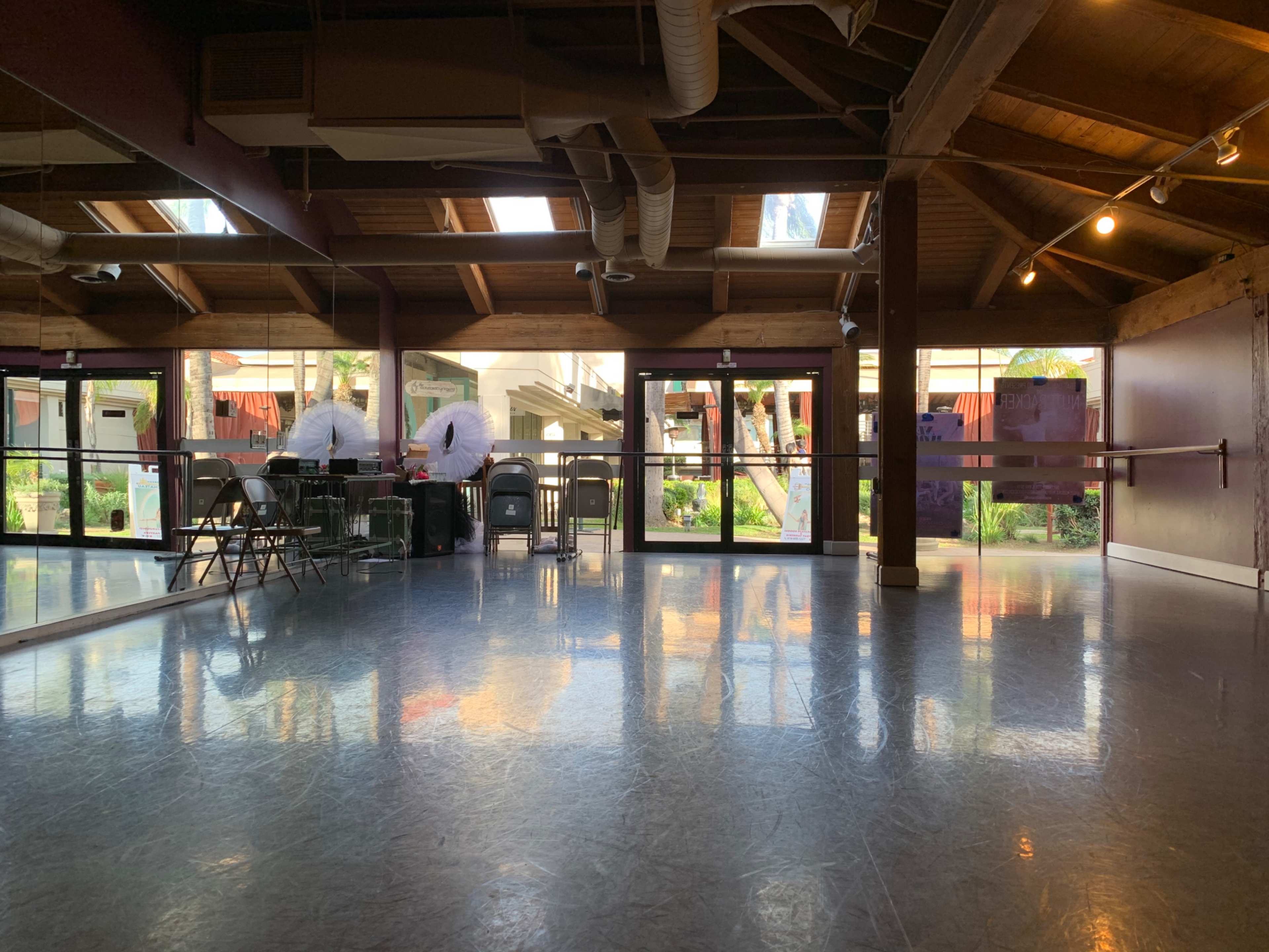 A dance studio with wooden beams and a mirrored wall, featuring empty chairs and large windows that overlook an outdoor area.