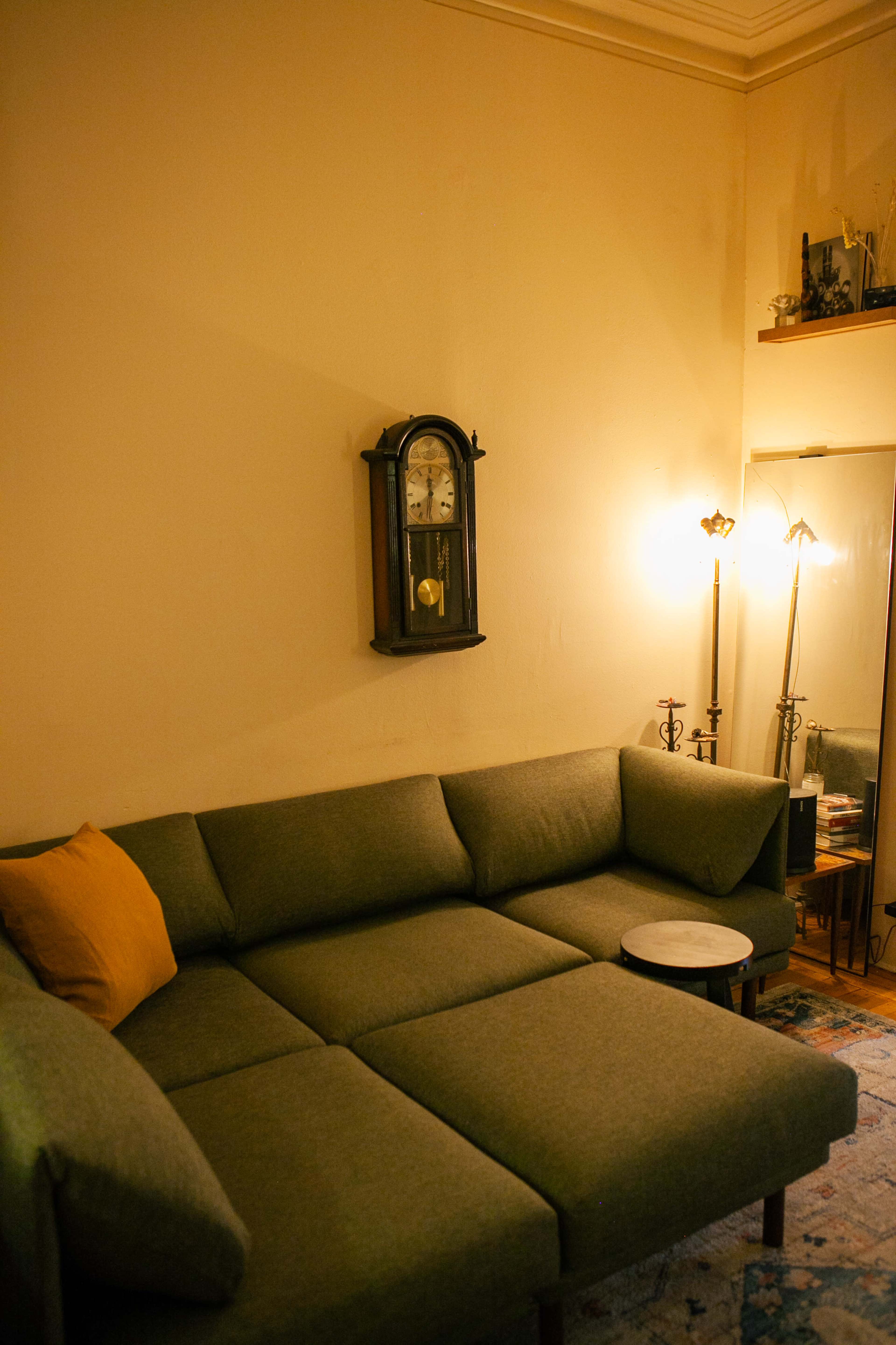 The image shows a green sectional sofa in a room with a wall clock, a round coffee table, and floor lamps.