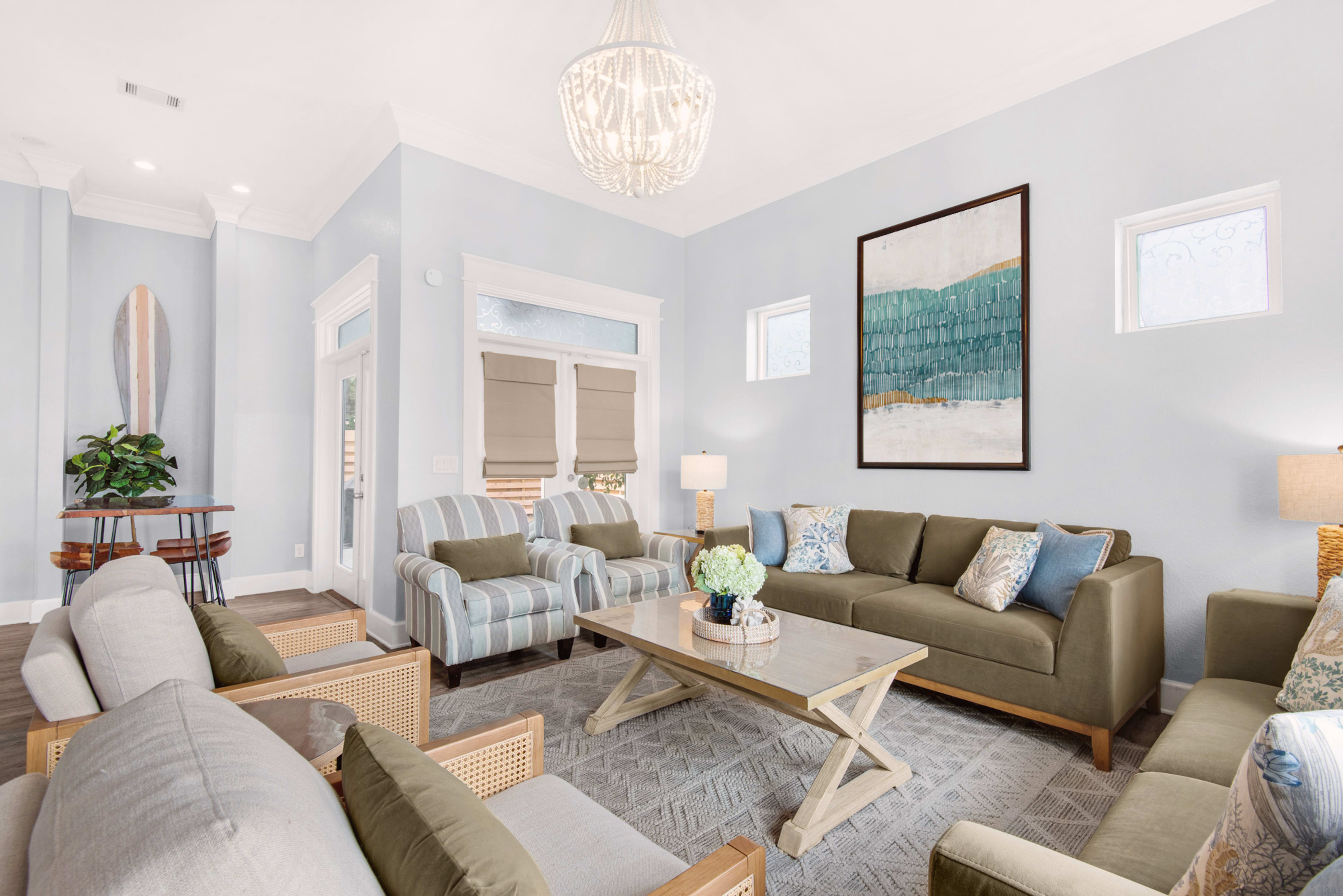 The image shows a modern living room with a high ceiling, featuring a large chandelier, a coffee table, a mix of seating options, and a decorative wall art piece.