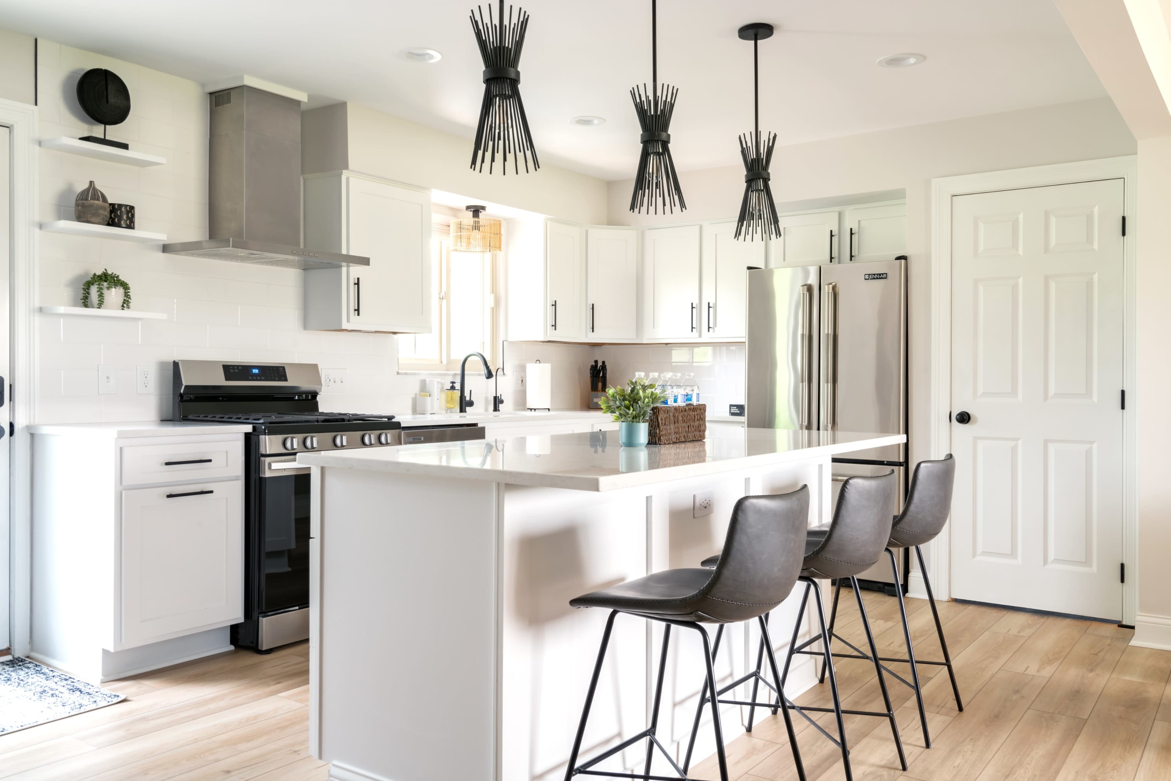 A modern kitchen with a central island, three bar stools, and pendant lighting above, featuring white cabinetry and stainless steel appliances.