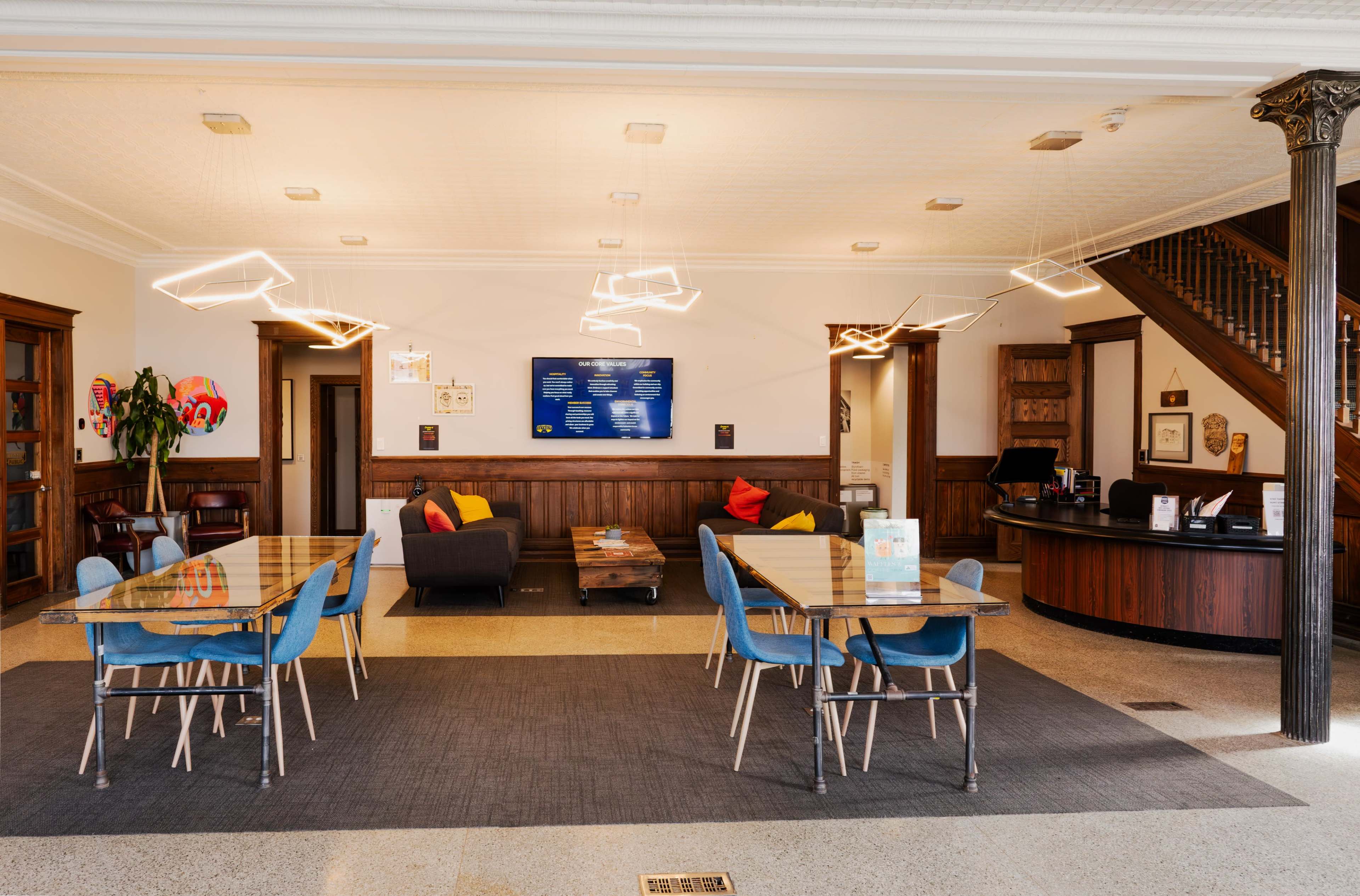 A modern reception area with seating, a front desk, and tables arranged on a carpeted floor beneath stylish light fixtures.