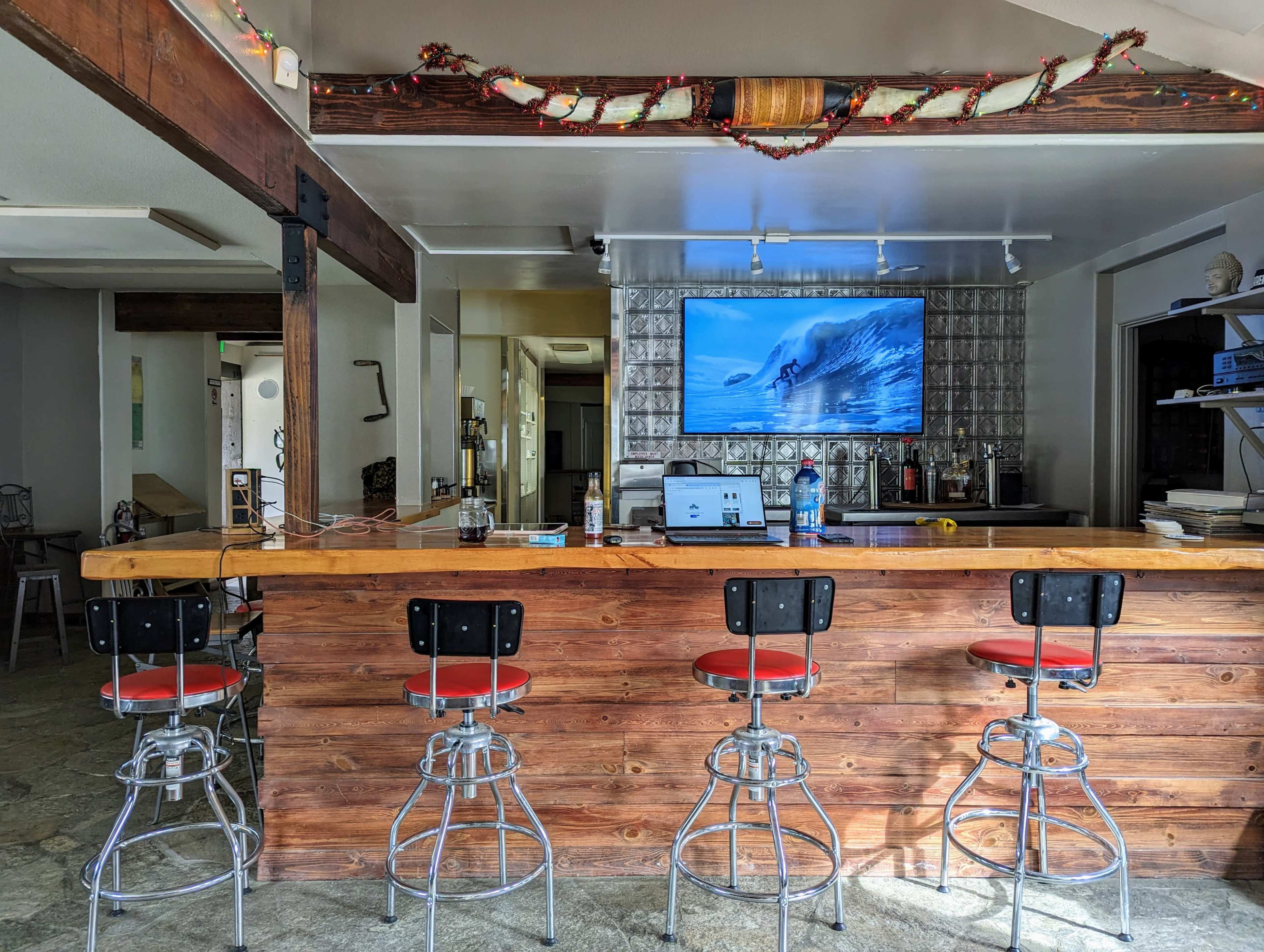 A wooden bar with four high stools is coupled with a large television displaying a surfing scene, set in a spacious interior with decorative garlands.