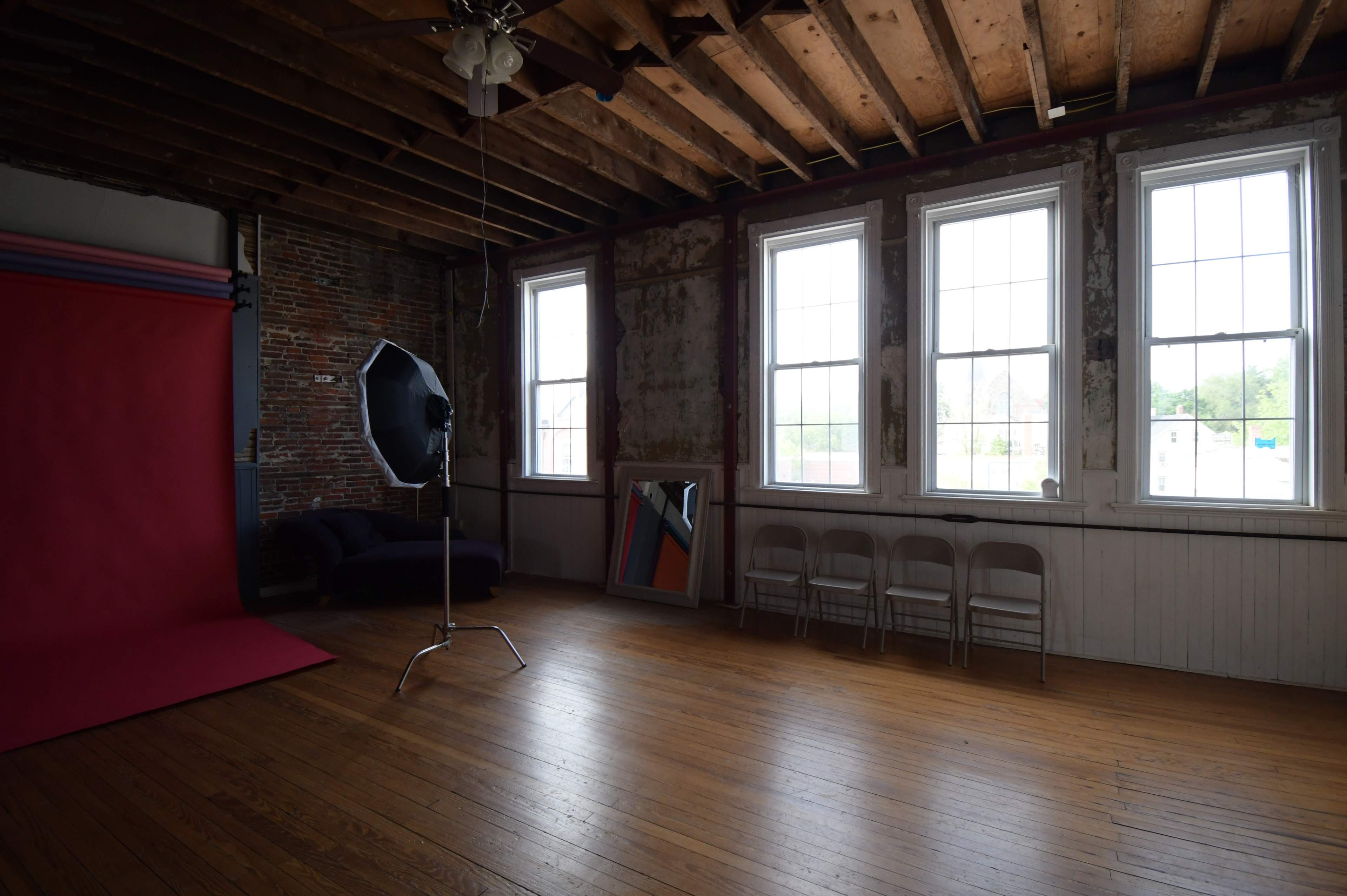 A spacious photo studio with a red backdrop, three tall windows, a mirror, and several white chairs arranged along the wall.