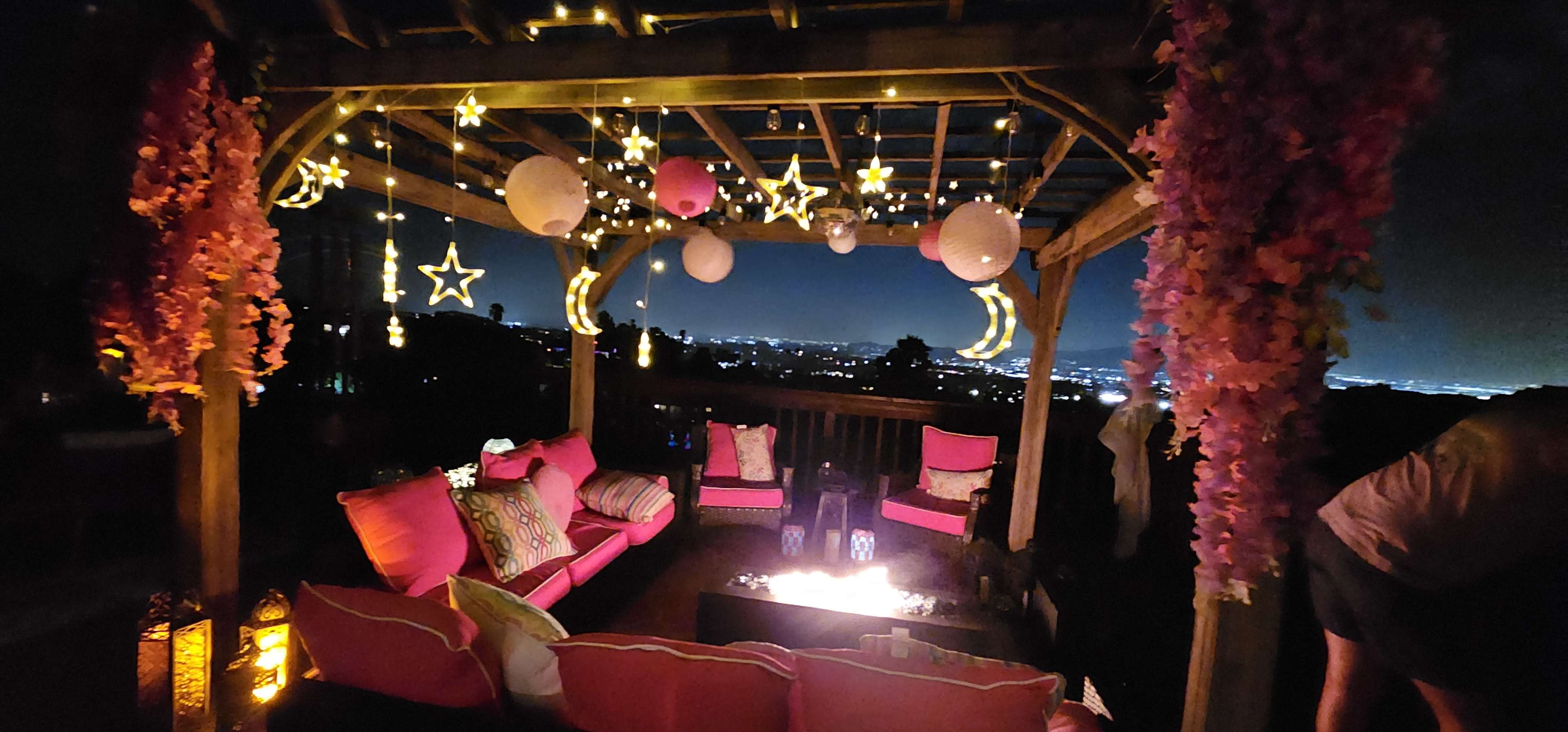A decorated pergola with pink seating and string lights overlooks a city skyline at night.