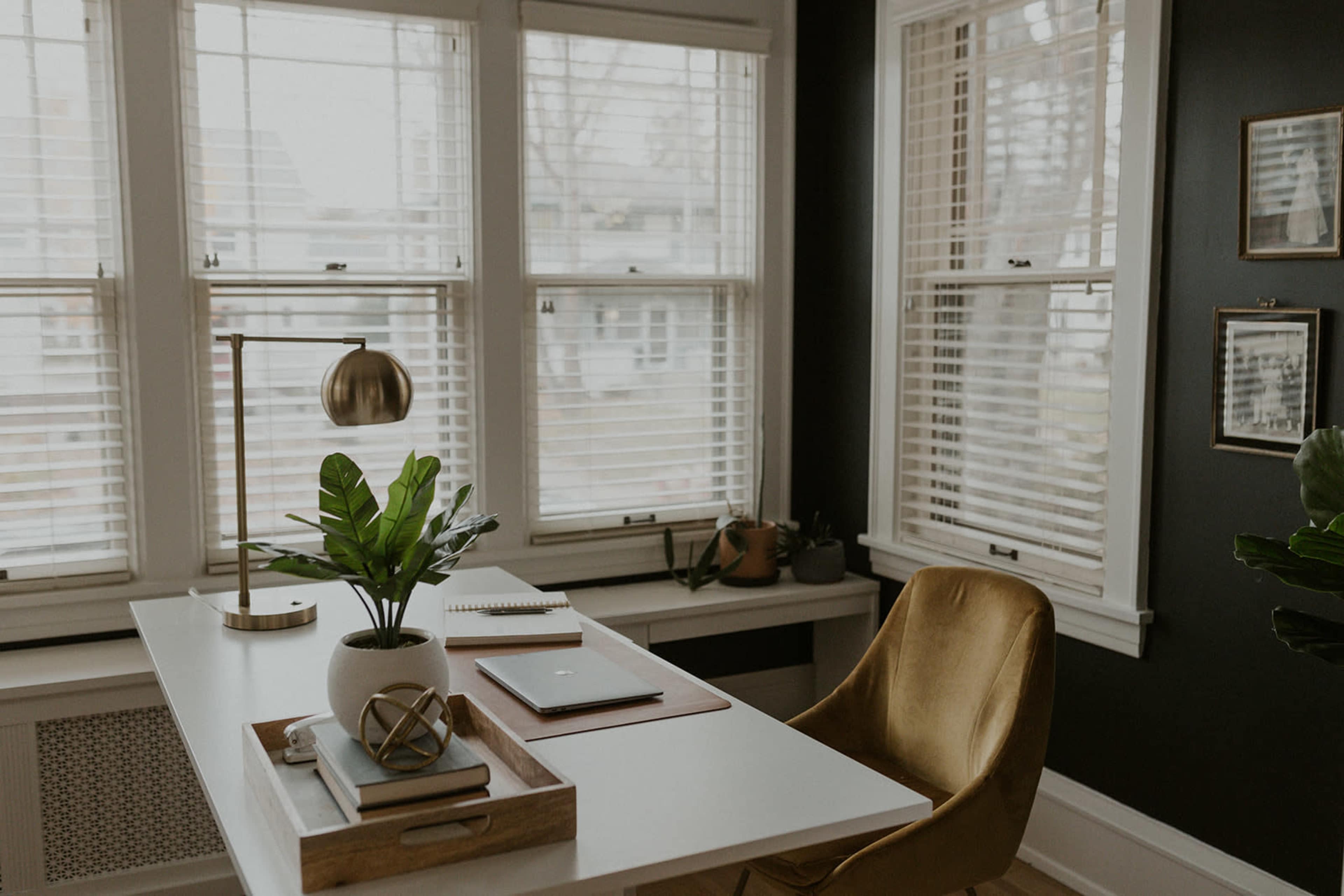 A modern office space features a white desk with a laptop, notebook, and decorative items, accompanied by a gold desk lamp and a green plant, set against large windows with blinds.