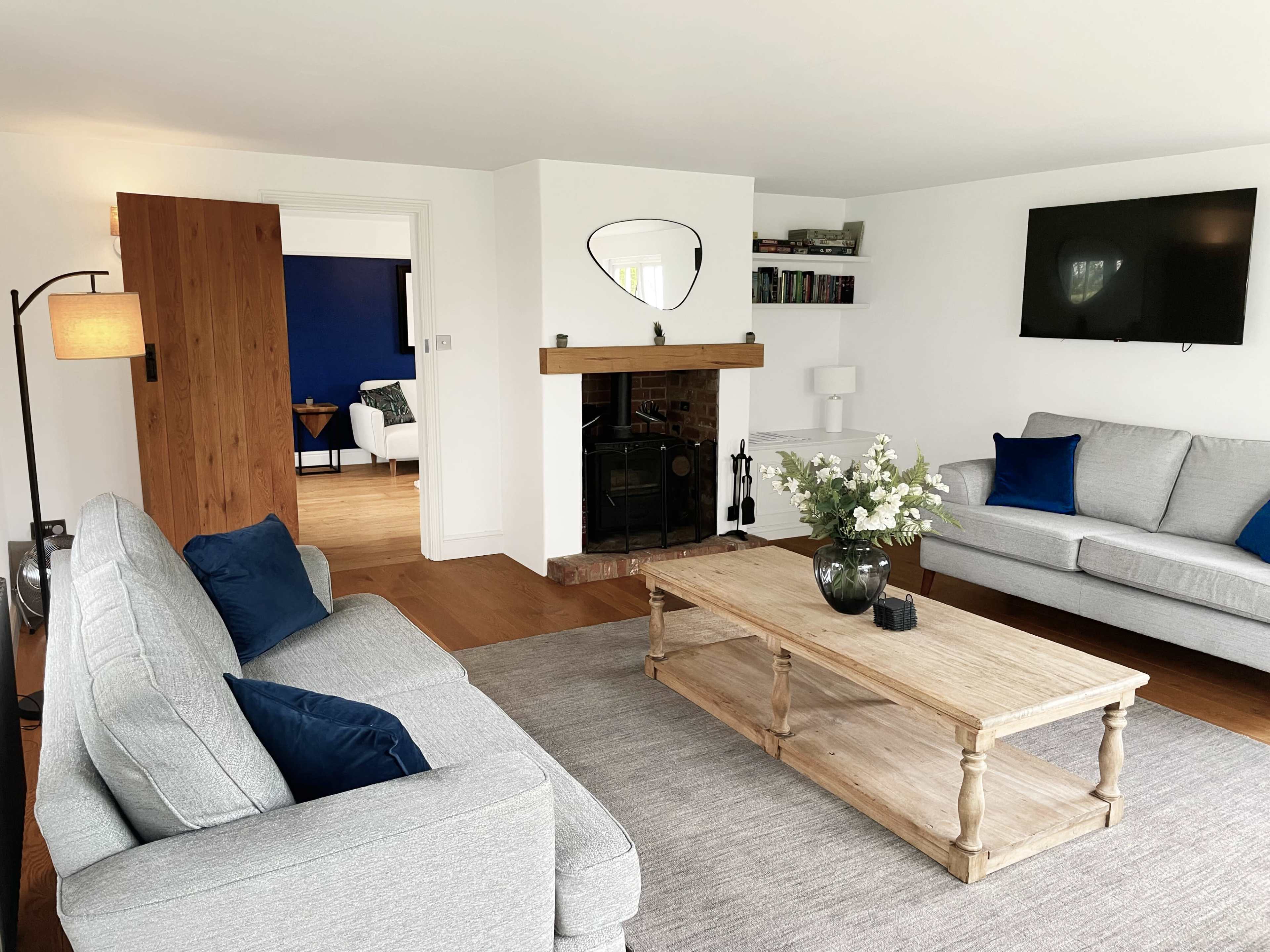 The living room features a light gray sofa and a wooden coffee table, with a fireplace and a wall-mounted TV, set against a backdrop of white walls and hardwood flooring.