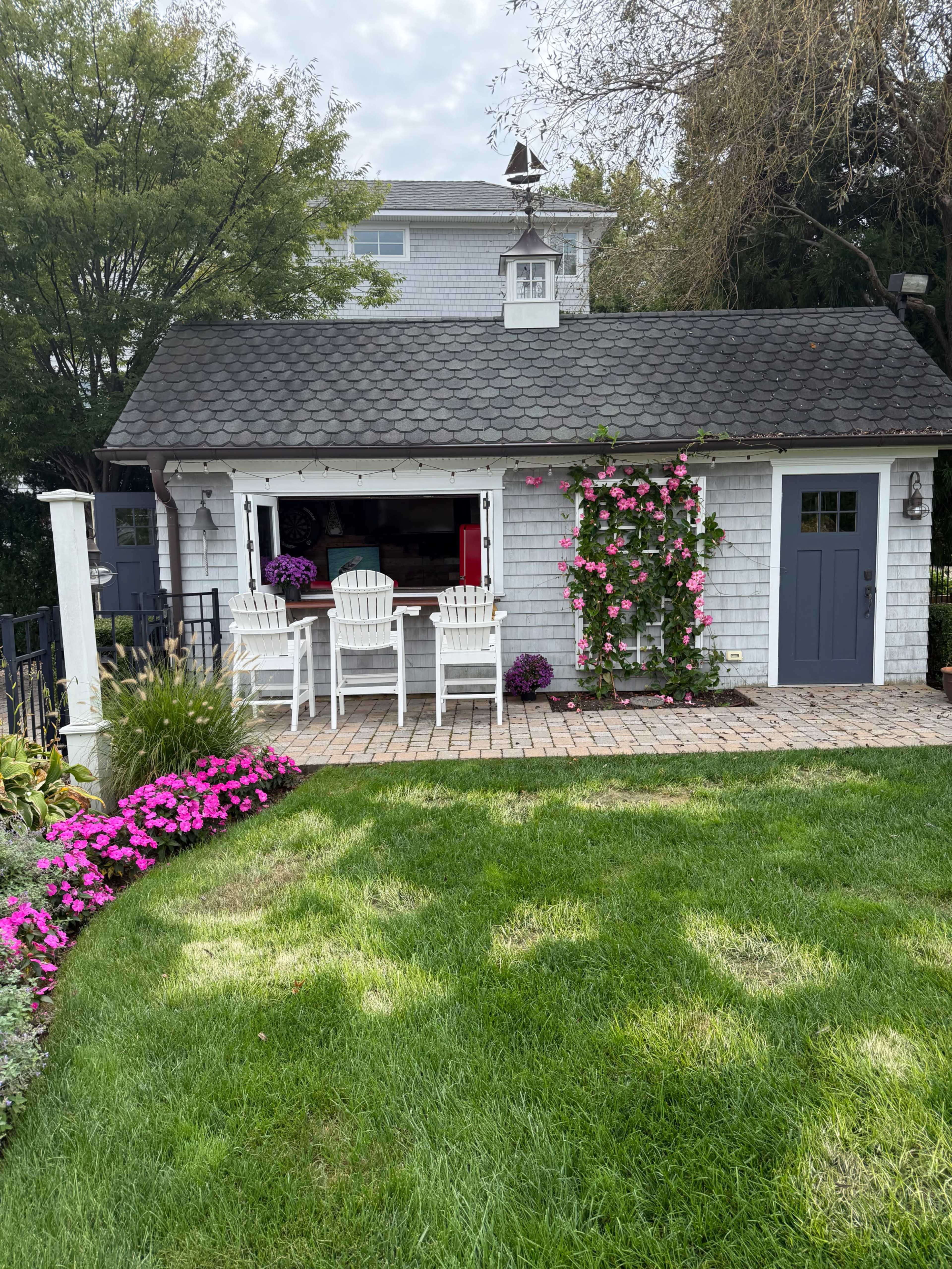 The image shows a charming cottage with a small patio area featuring white chairs, surrounded by colorful flowers and greenery.