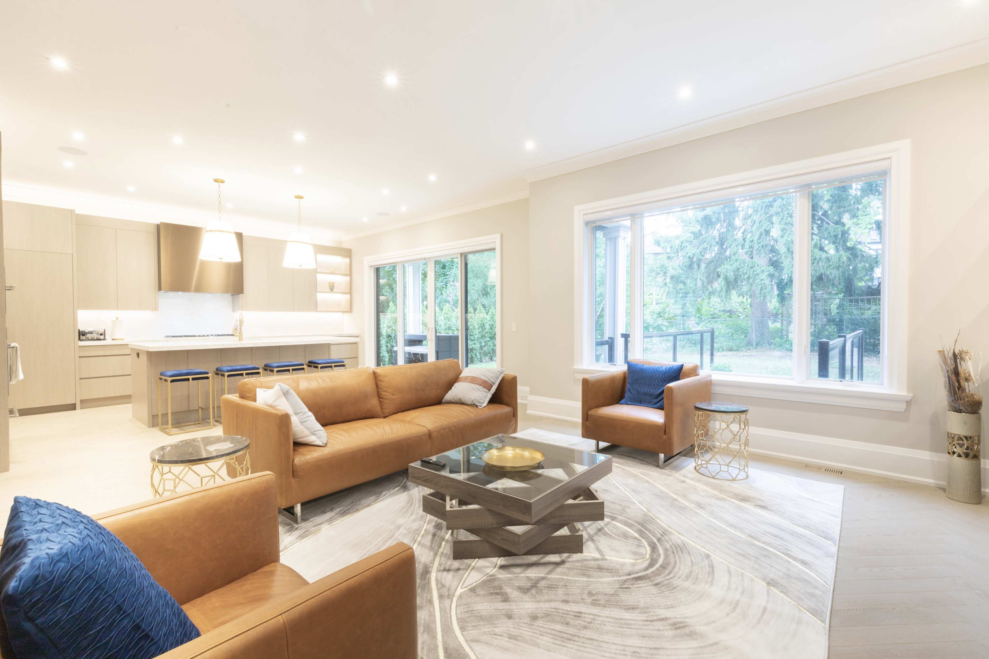 The living room features a brown leather sofa and armchairs, a coffee table, and large windows overlooking a backyard with trees.