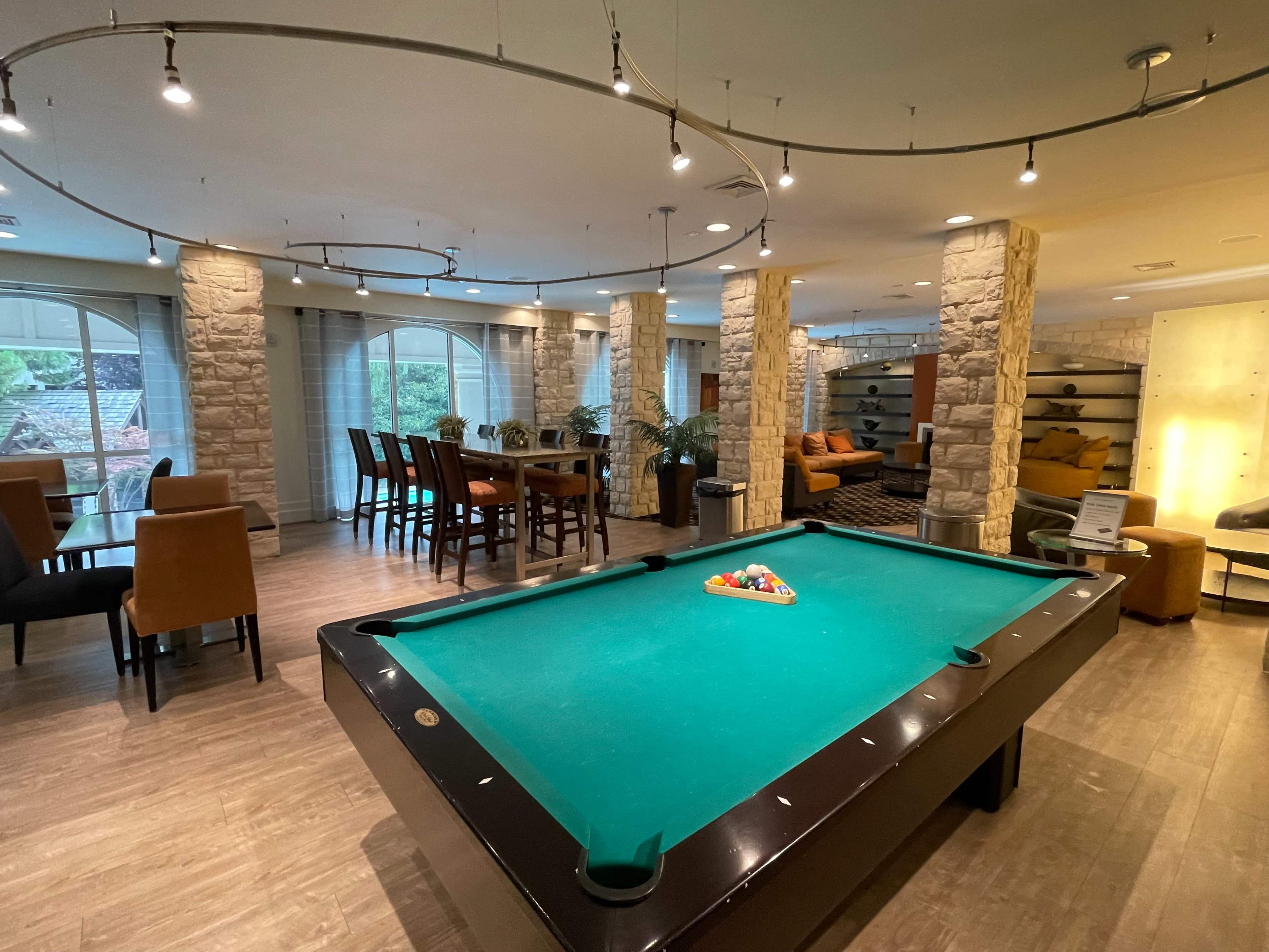 A spacious room features a billiards table at the center, surrounded by seating areas and dining spaces with modern decor.