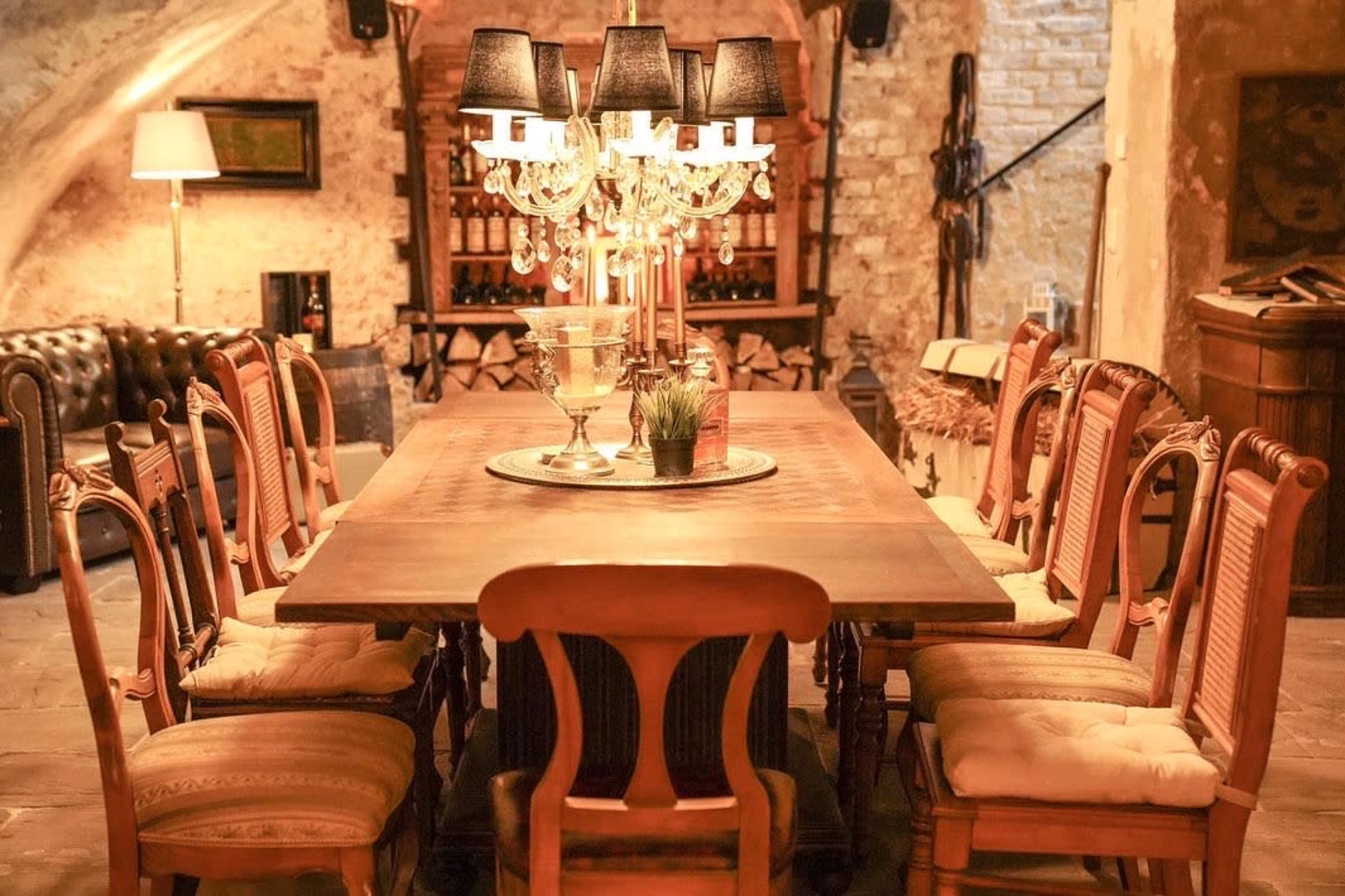 A large wooden dining table surrounded by eight ornate chairs sits under a chandelier in a rustic room with stone walls.