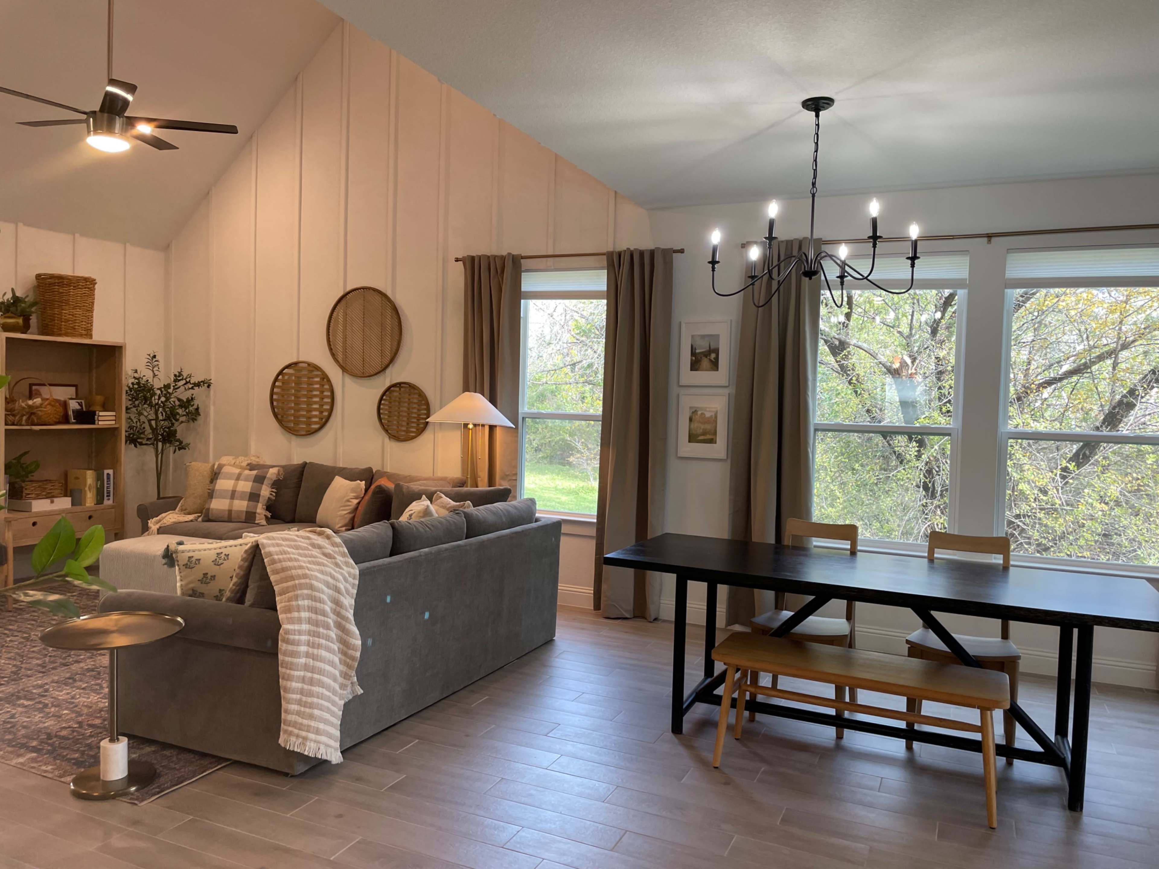 The image shows a cozy living space featuring a gray sofa with decorative pillows, a wooden dining table, a chandelier, and large windows with a view of greenery.
