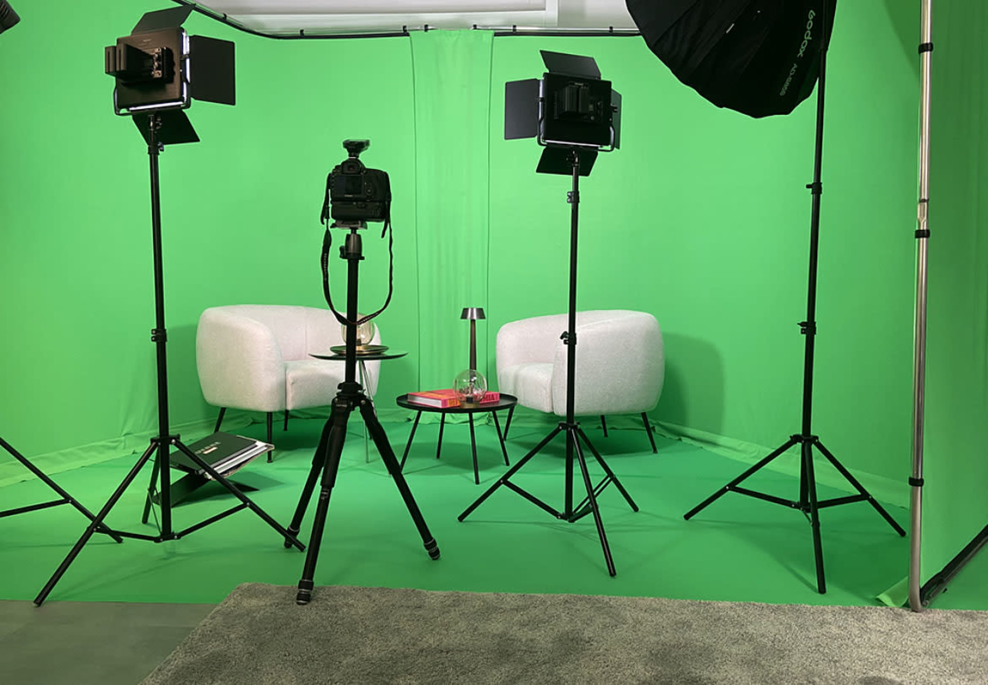 The scene shows a photo studio with two white chairs, a small round table, and a green backdrop, along with lighting equipment and a camera on a tripod.